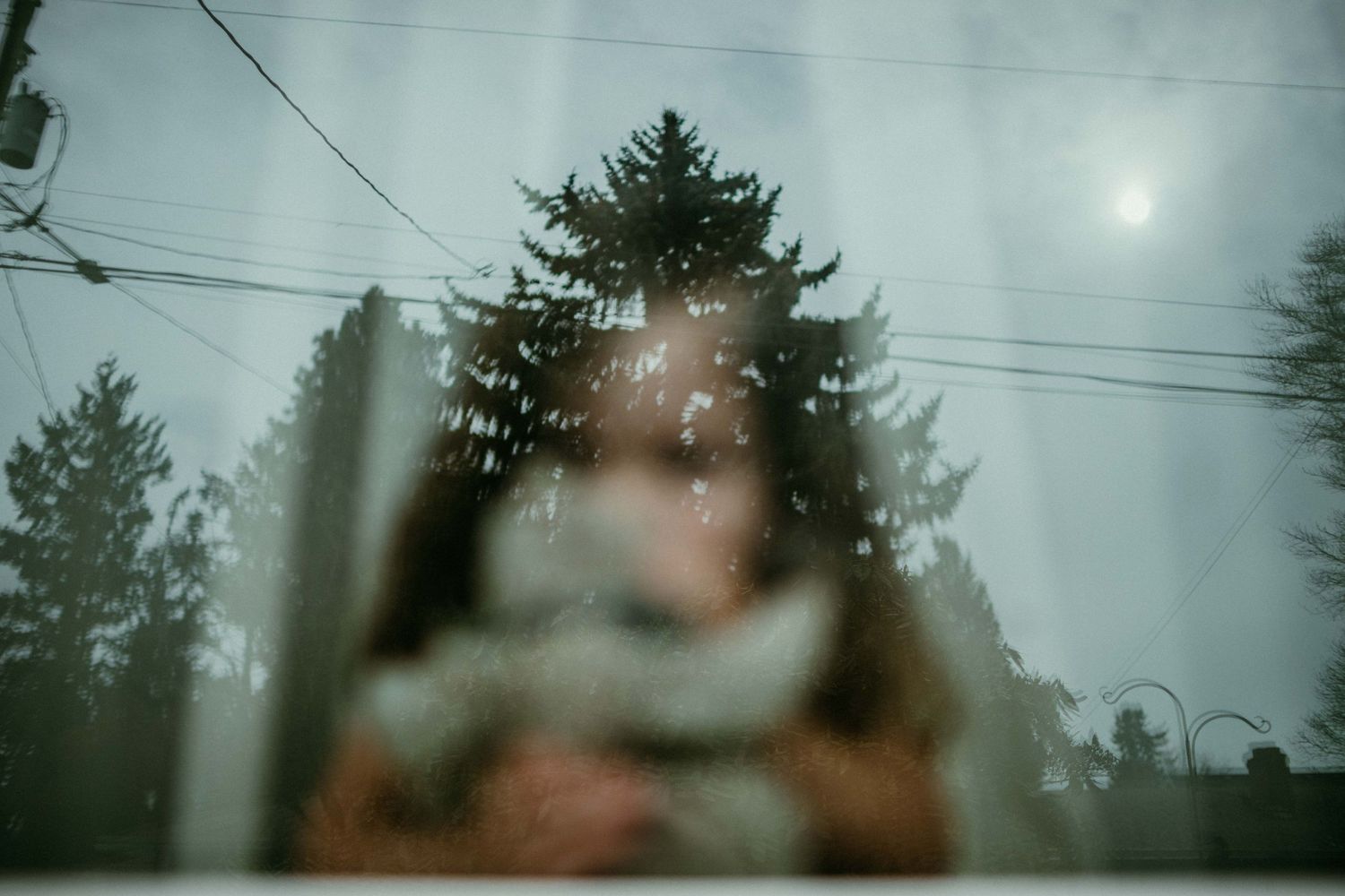 out of focus child looking through window at camera. reflection of tree and sun are in focus. portland family photog