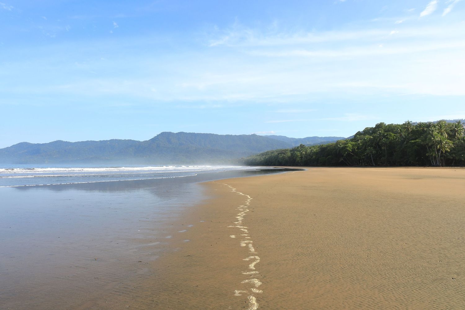 Pristine sandy beach stretches along coastline with mountain backdrop and gentle waves.