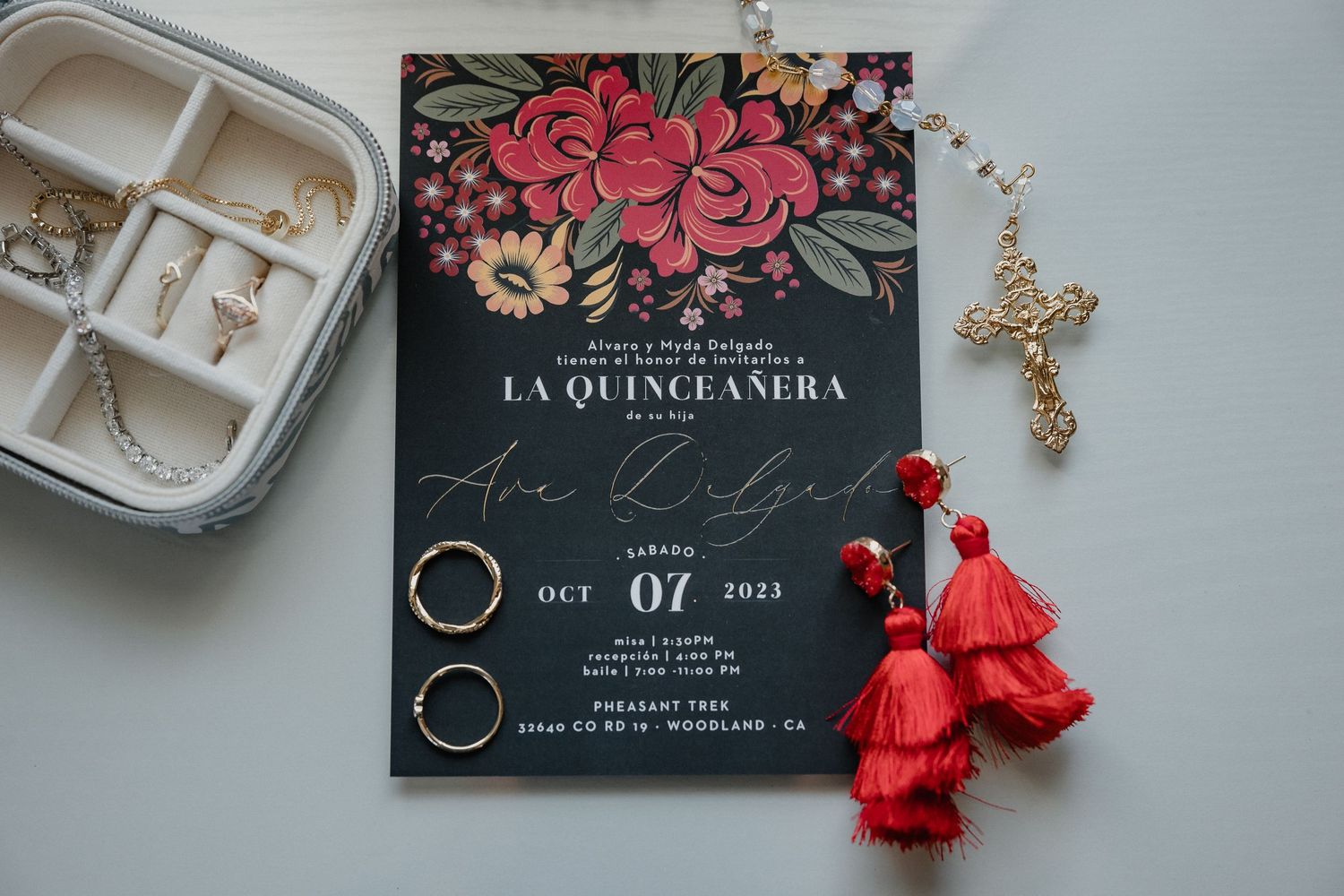 Quinceañera invitation with red floral design next to jewelry and tassels.