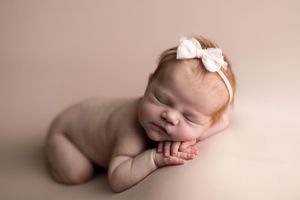 newborn baby girl with red hair laying on belly with head on hands on pink background with pink bow