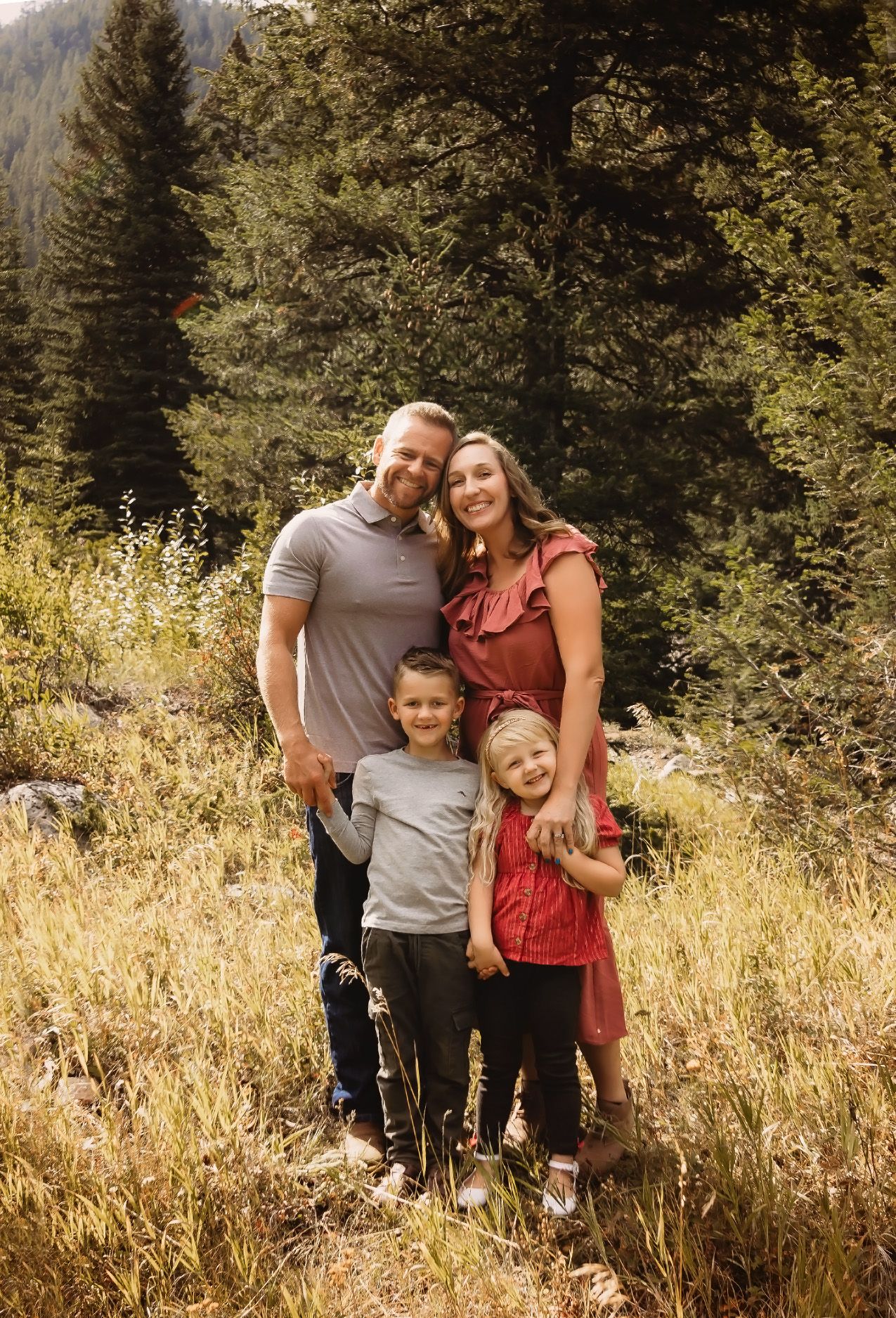 A family poses together in a sunlit meadow with evergreen trees in the background on a warm autumn day.