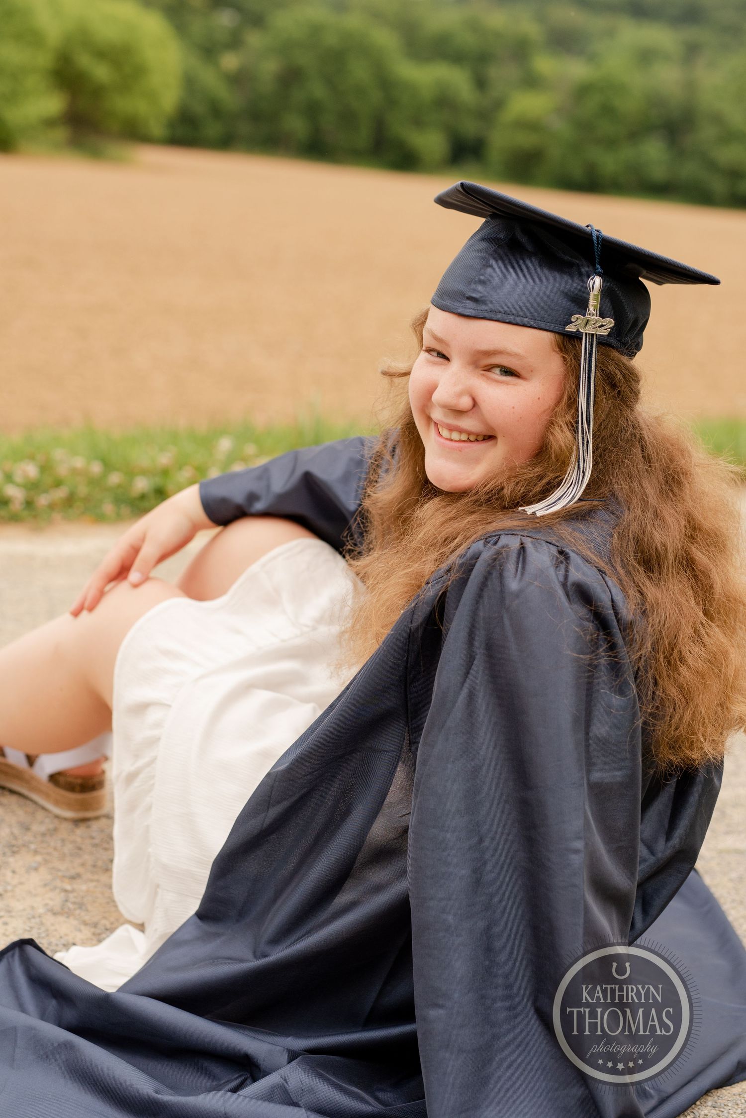 Graduate in academic regalia poses outdoors with rolling hills in background.