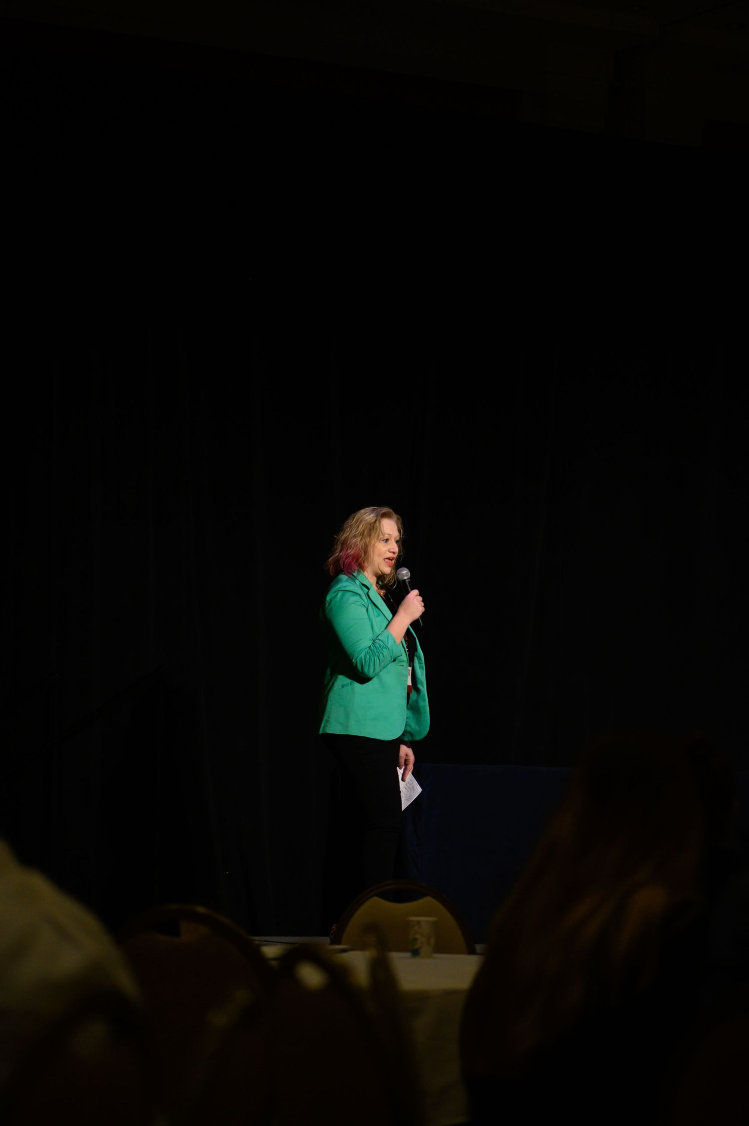 Performer in green blazer stands spotlit on dark stage.
