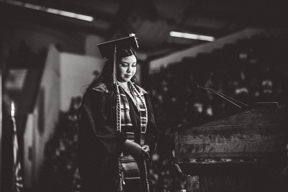 Graduate in traditional cap and gown gives speech in dramatic black and white photo.