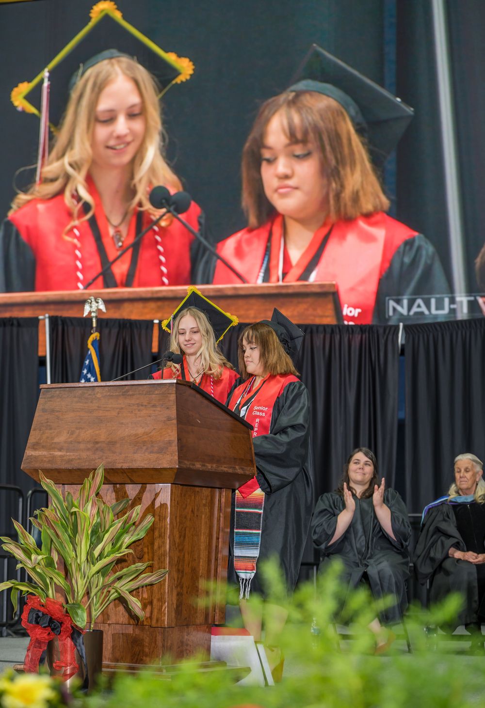 Two graduates in red regalia speaking at podium during commencement ceremony.