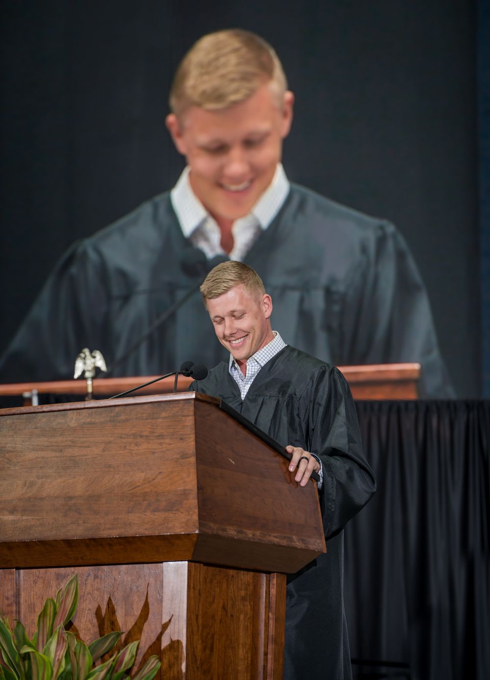Speaker at a podium delivers remarks during a graduation ceremony with their portrait displayed on screen behind them.