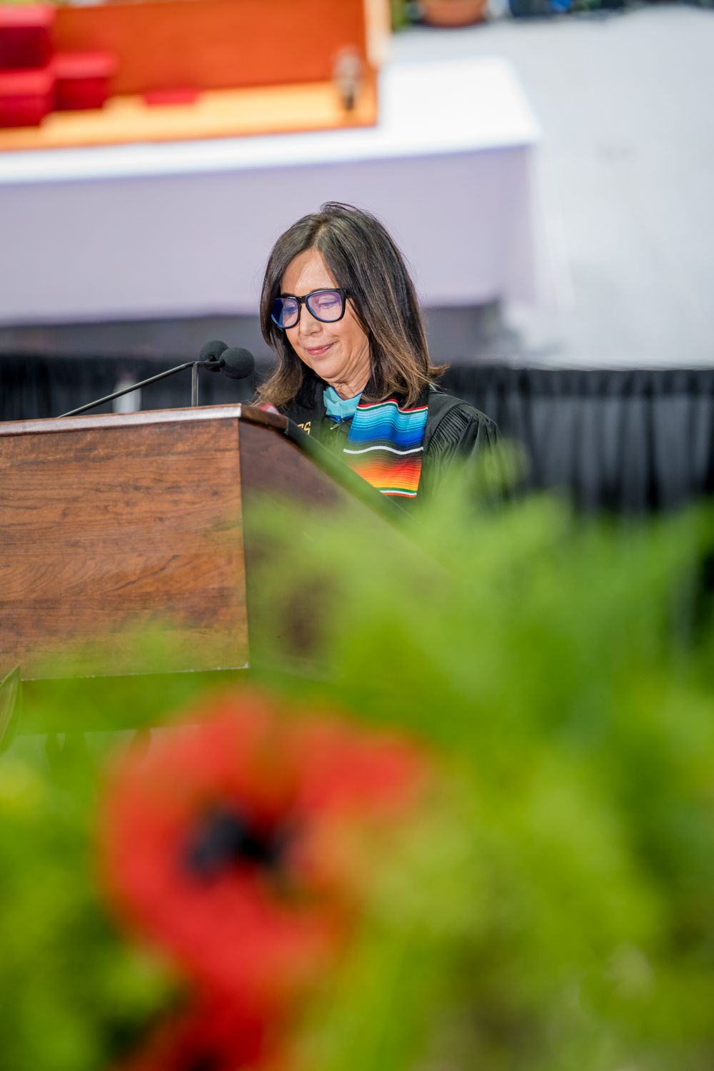Speaker in academic regalia smiles while giving remarks at a wooden podium with colorful flowers in foreground.