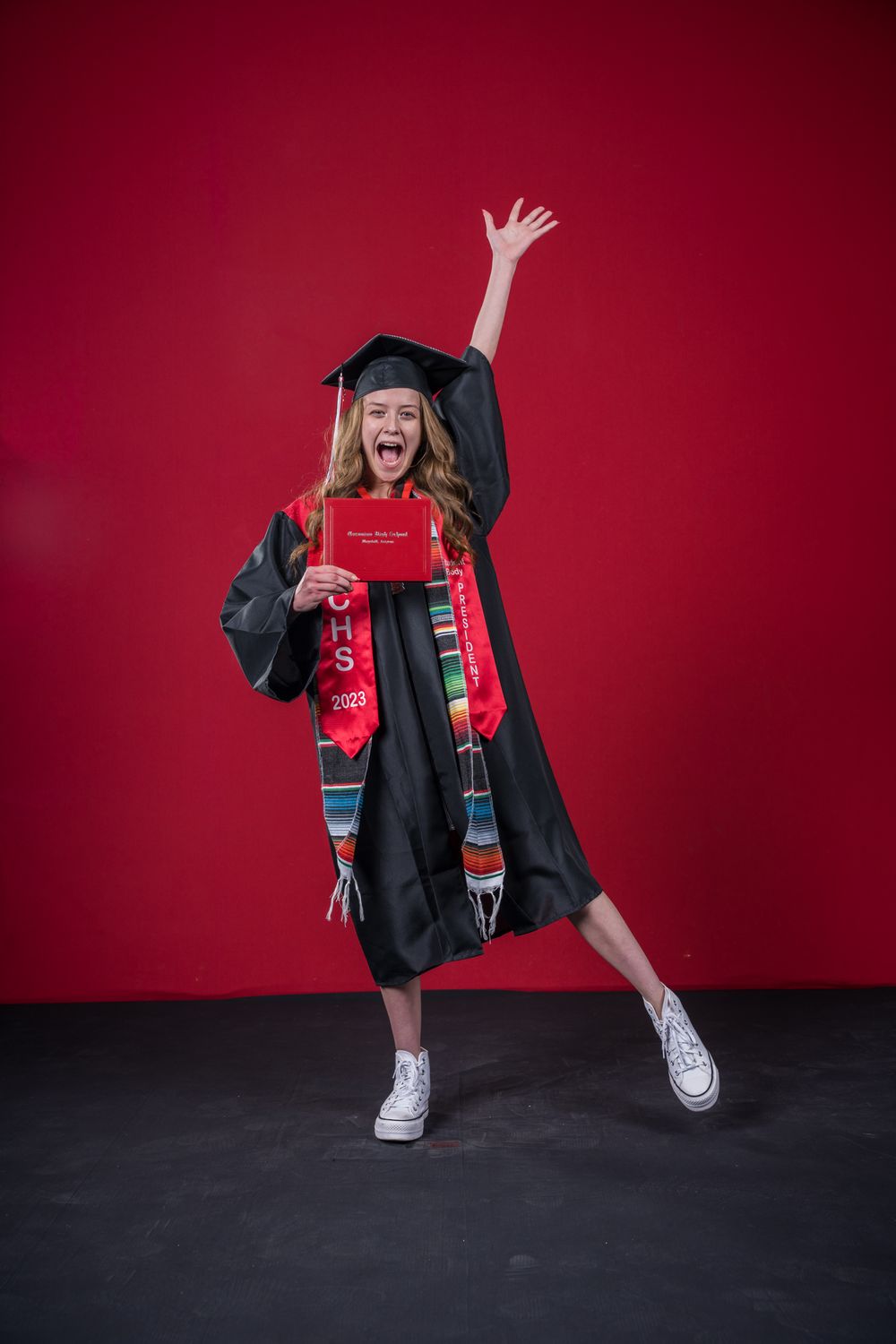 Graduate in cap and gown jumps joyfully while holding diploma against red background.