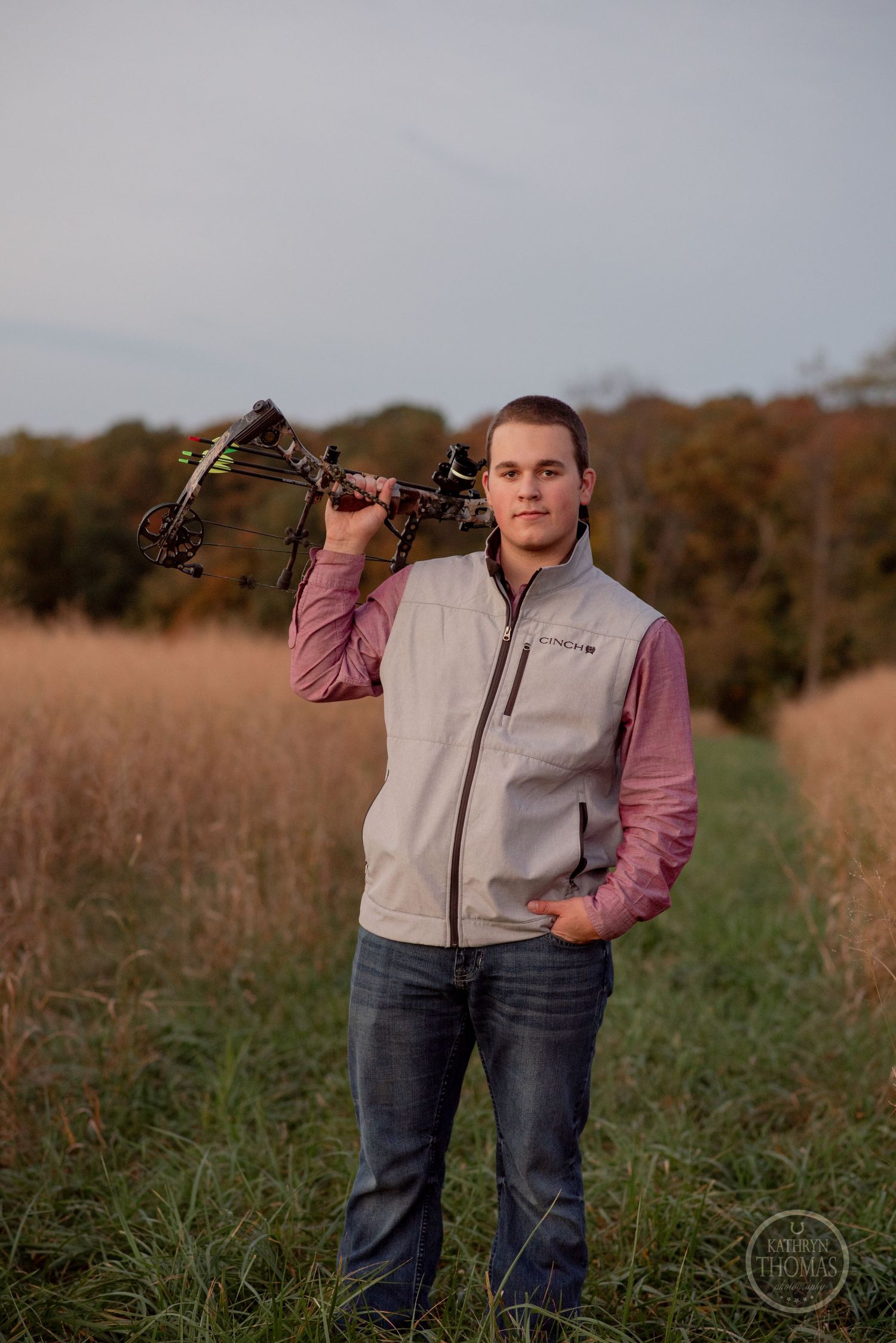 Person poses with compound bow while wearing gray vest and pink shirt in grassy field.