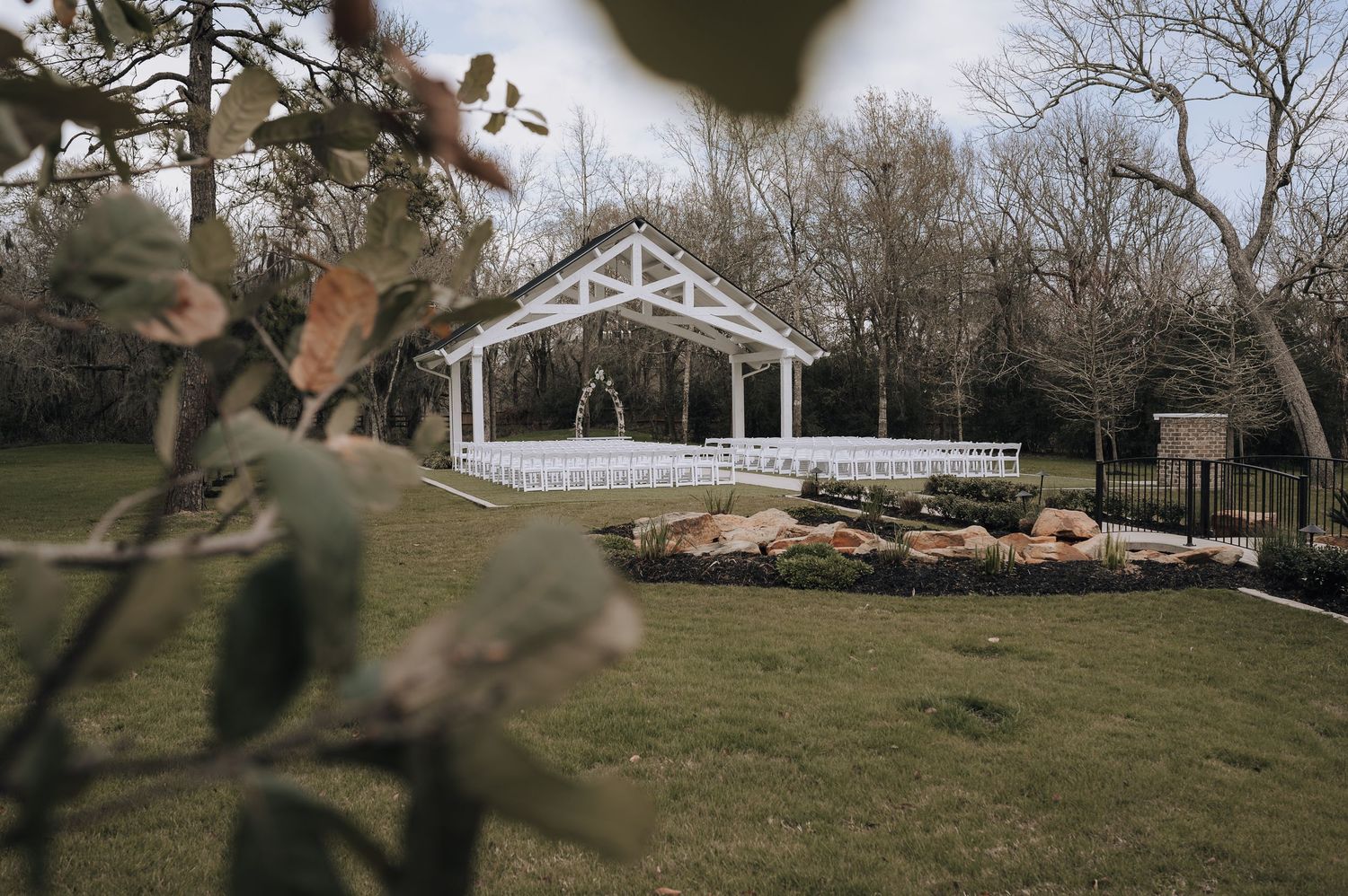 A white wooden wedding gazebo sits on landscaped grounds with decorative rocks and trees.