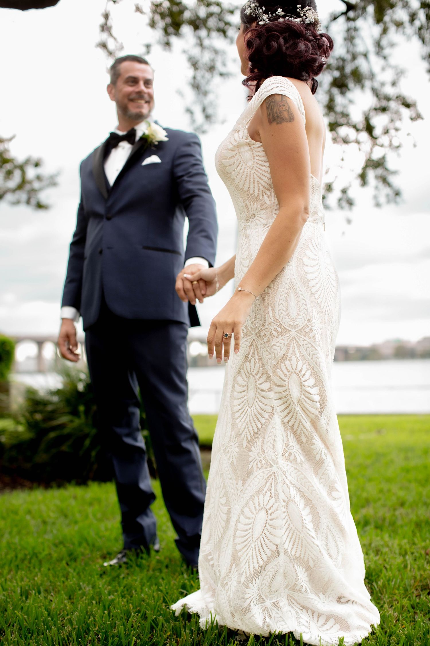 A wedding couple holding hands outdoors, with the bride wearing an ornate lace dress and the groom in a navy suit.