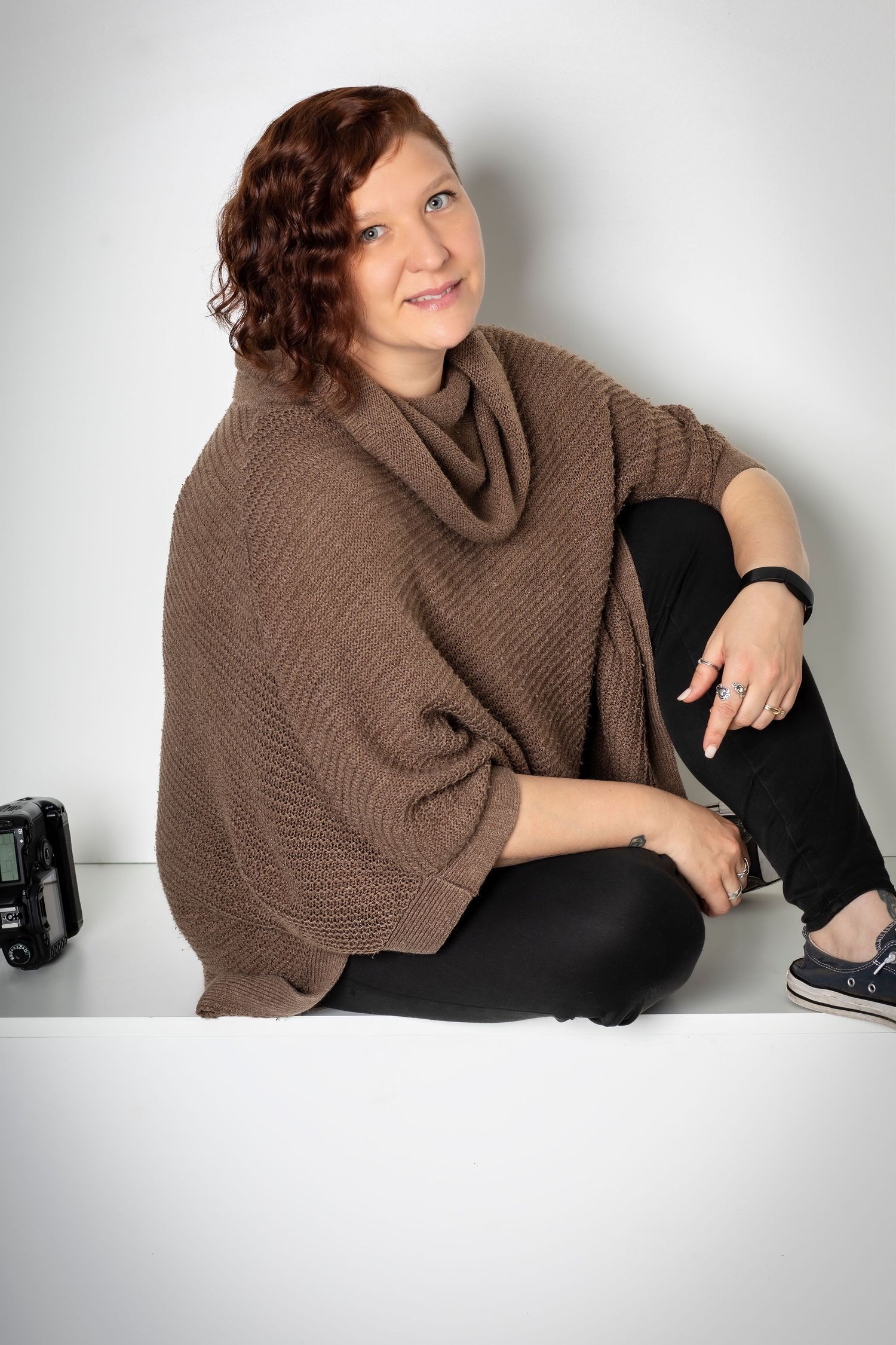 Portrait of photographer wearing brown poncho sweater sitting on white backdrop.