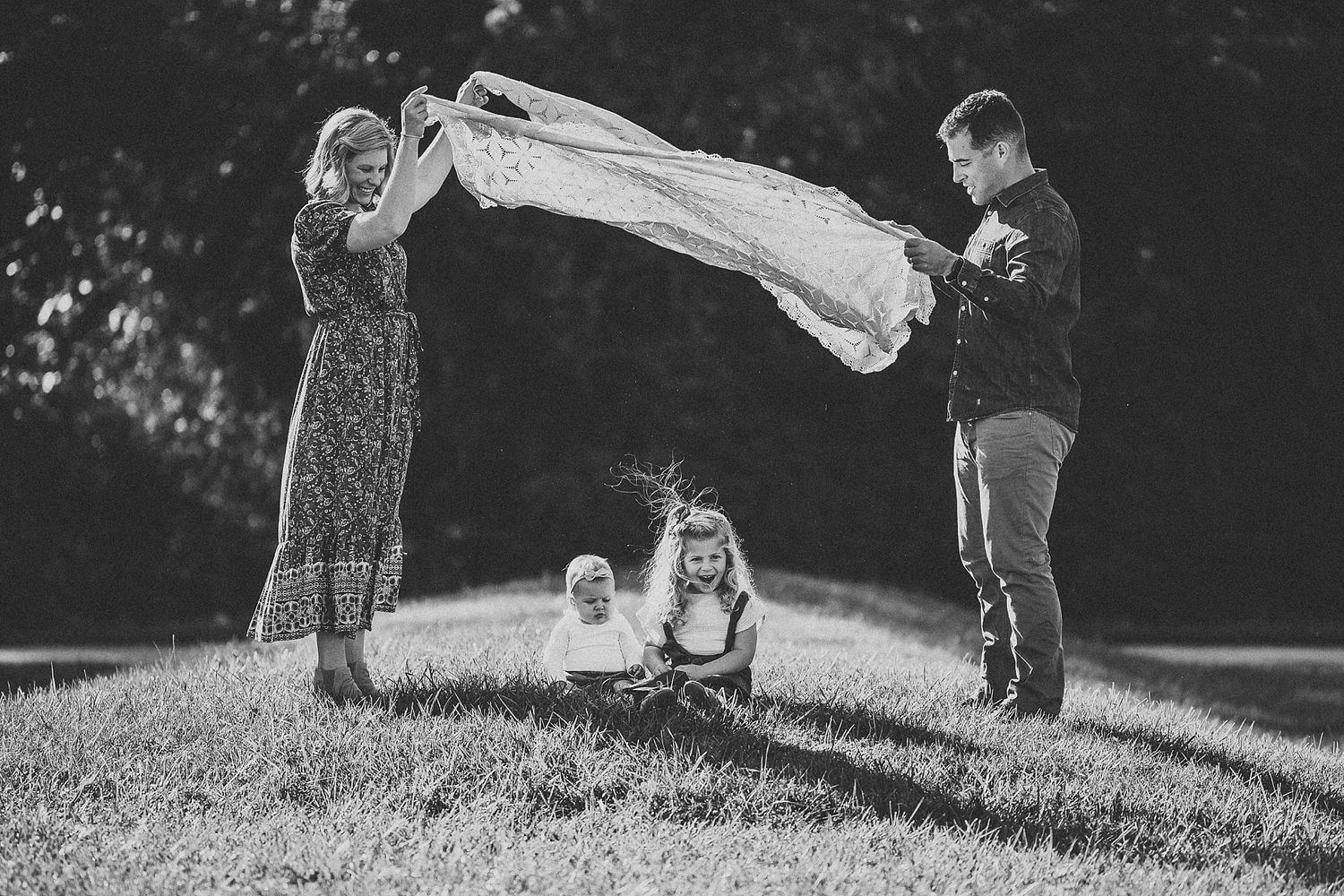 Black and white photo of family playing with blanket in grass outdoors.
