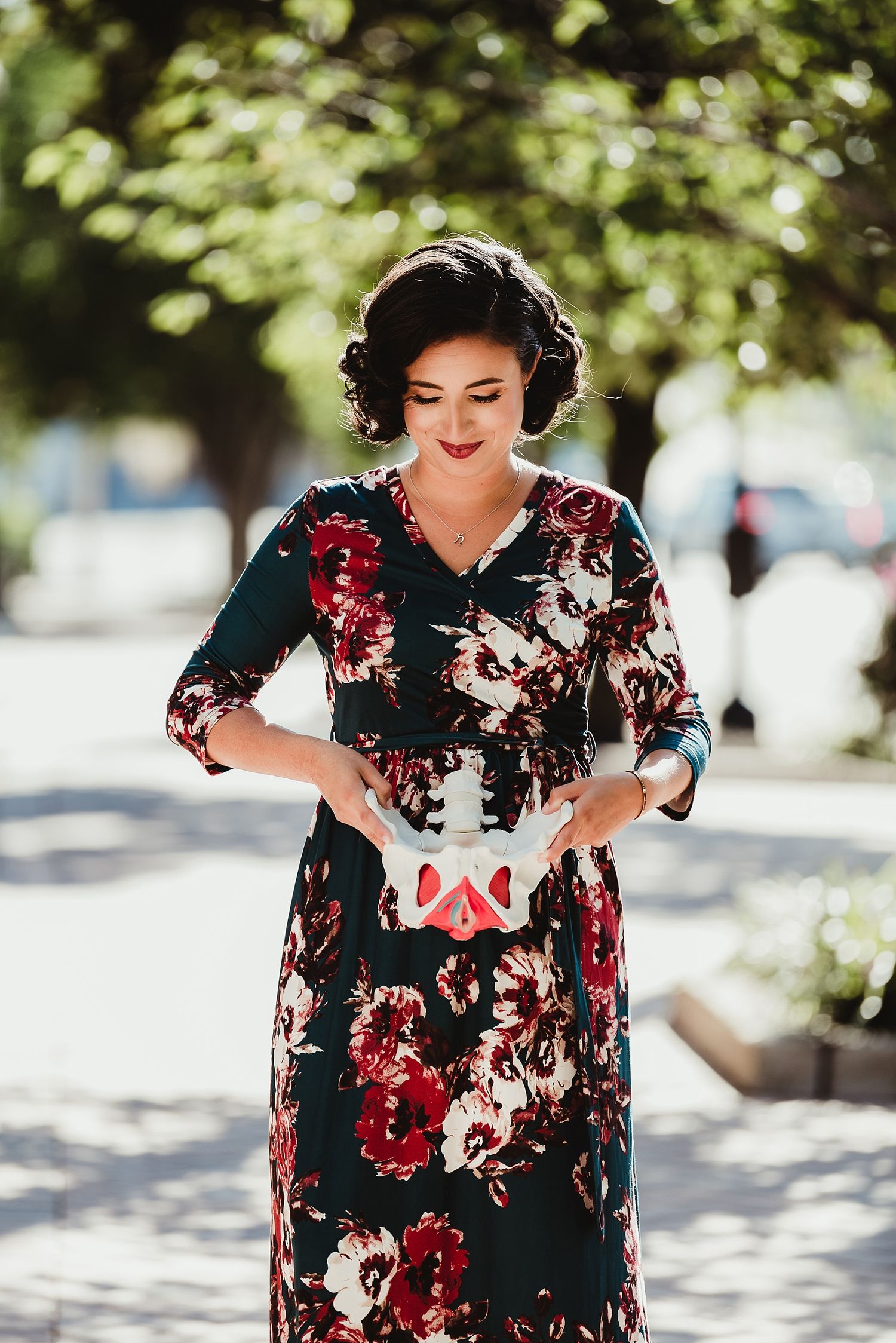 Woman in a vintage-style floral dress stands in dappled sunlight on a city sidewalk.