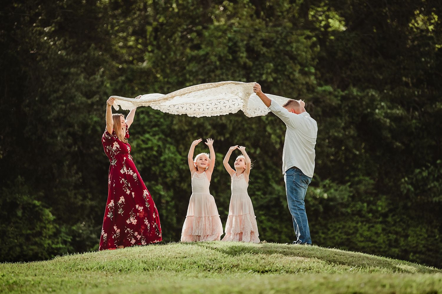 Family creates a playful tunnel with fabric on a grassy hill during outdoor photoshoot.