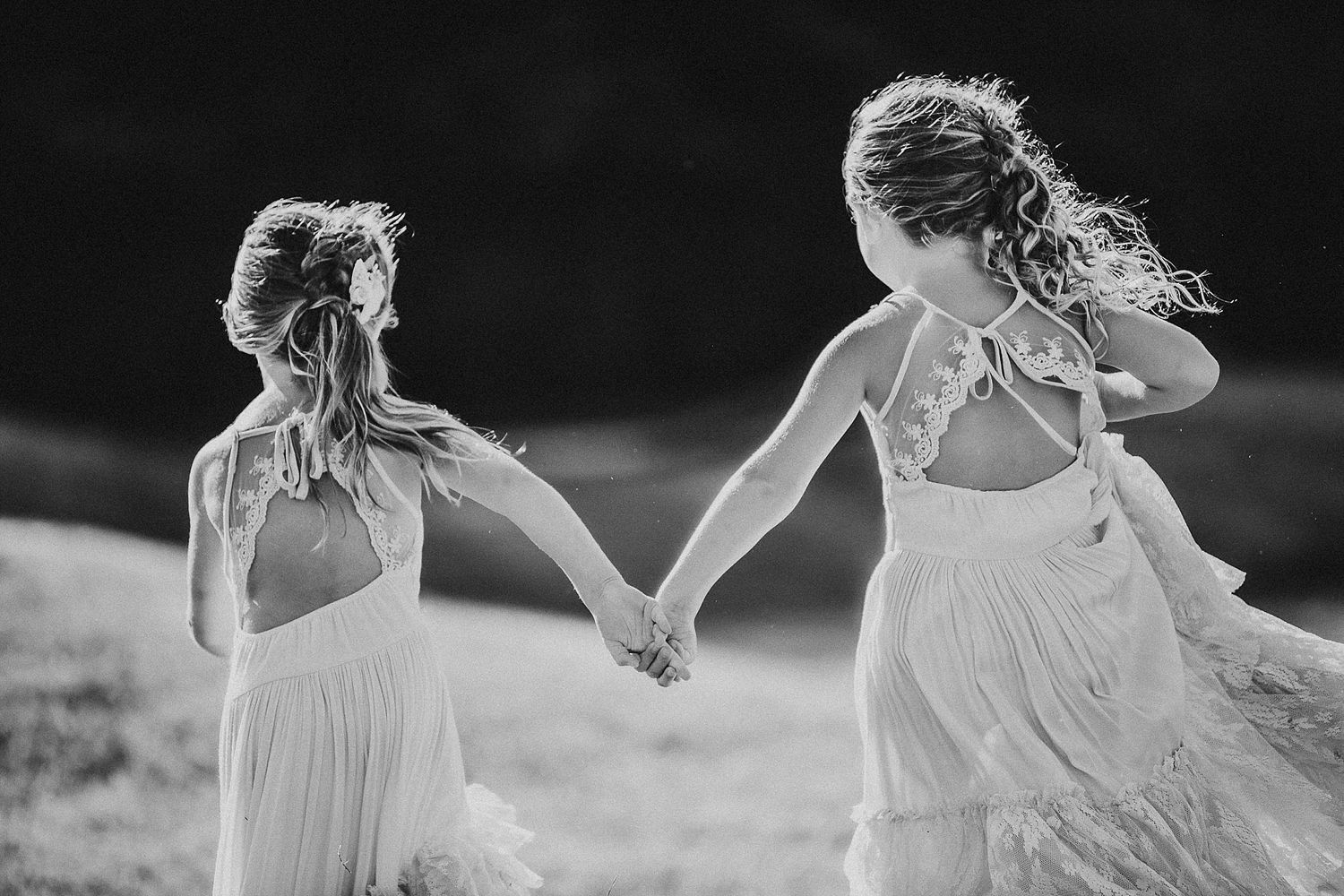 Two figures in flowing white dresses hold hands while running through a field.