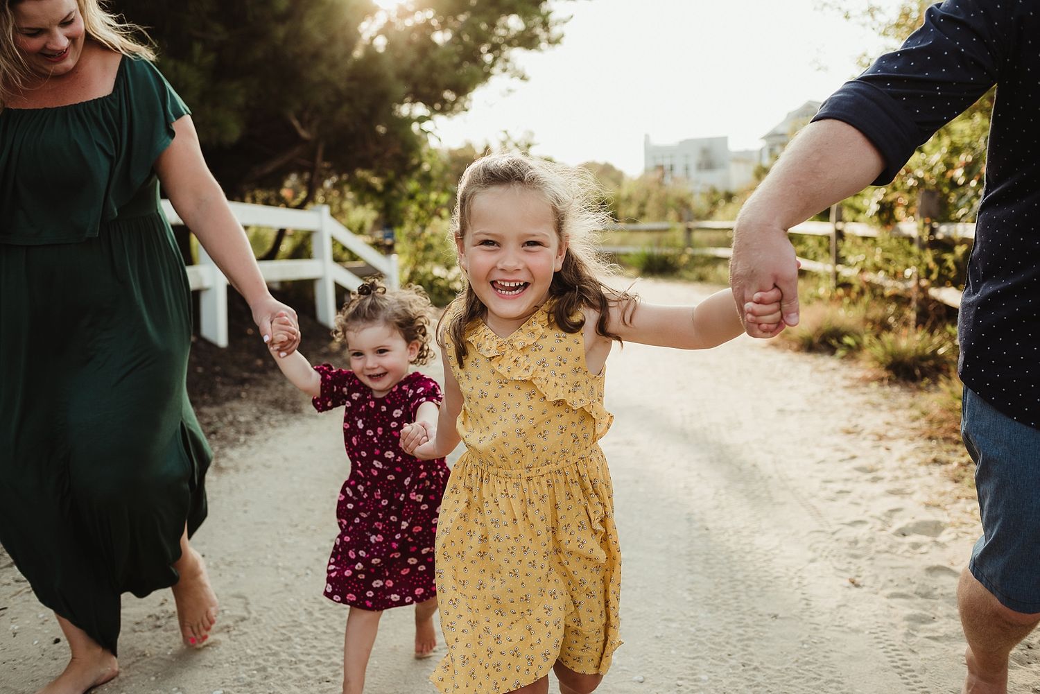 Children in floral dresses holding hands with their parents while walking on a sunny path.