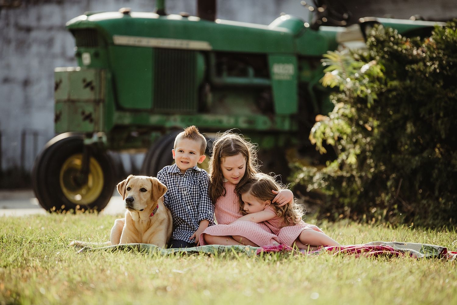 Children and a golden retriever sitting on a blanket with a green John Deere tractor in background.