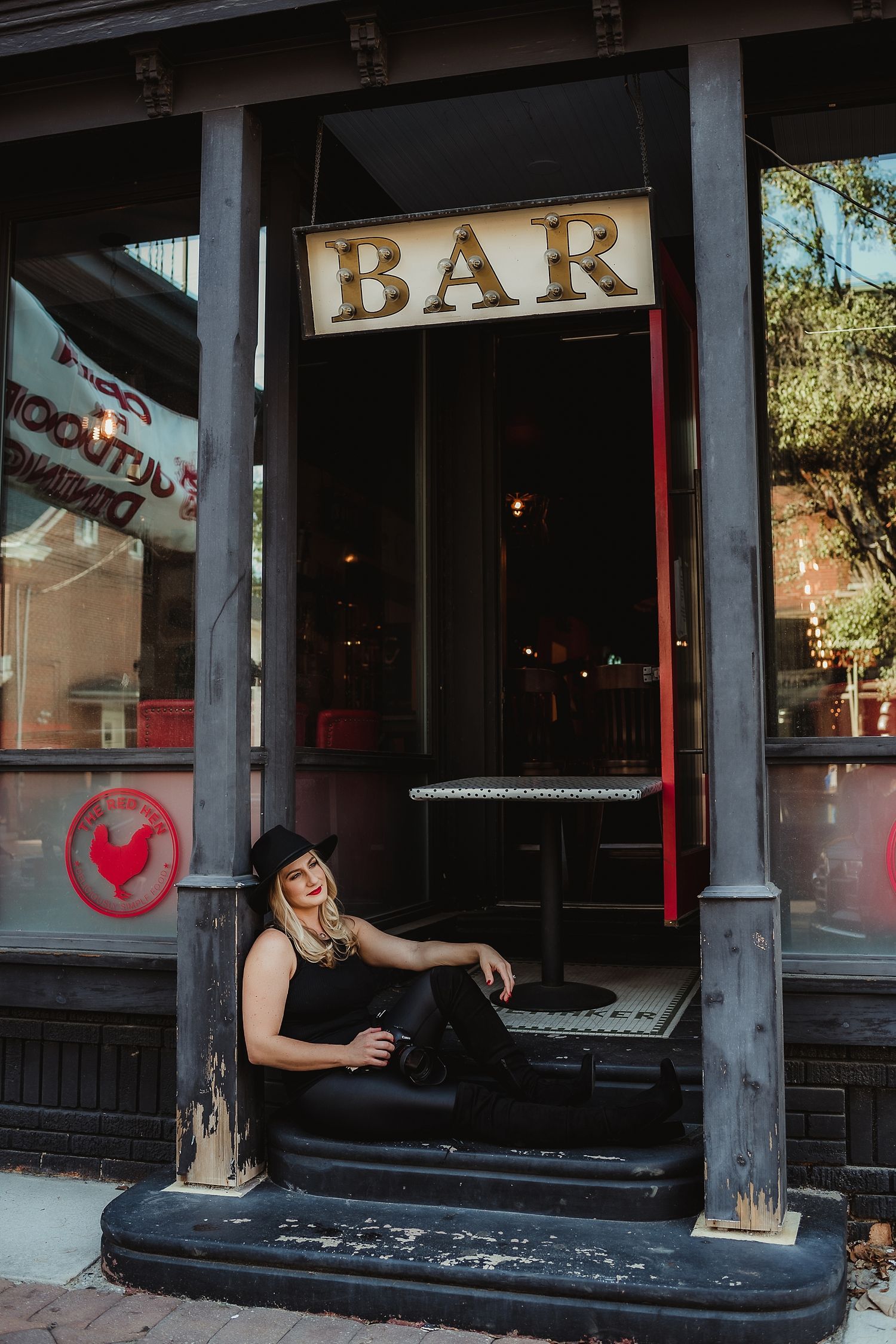A moody portrait of someone sitting on steps beneath a vintage bar sign.