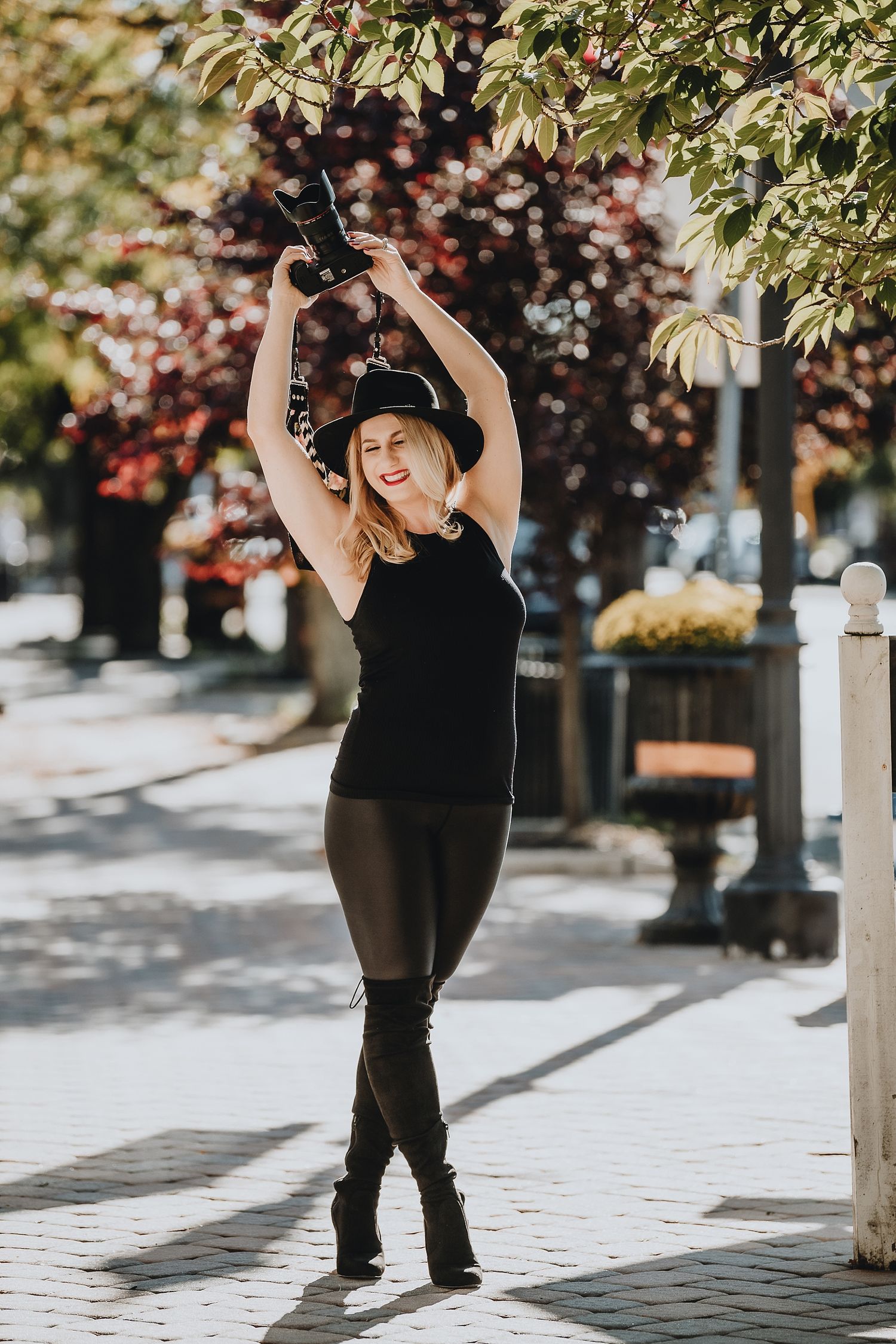 Woman in black outfit and hat stretching with camera in urban setting.