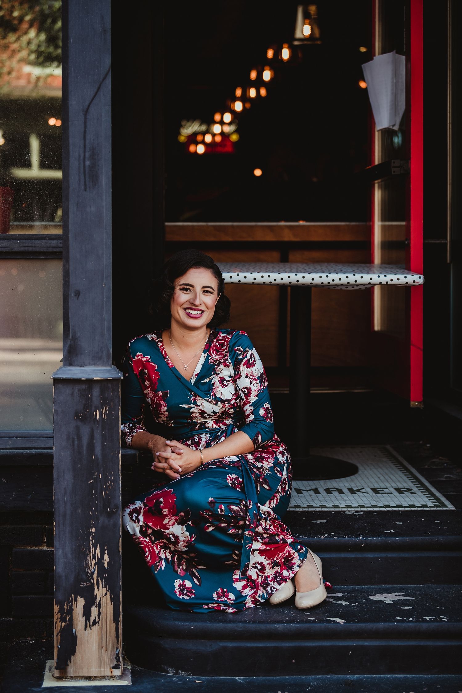 Woman in floral dress sitting on steps outside storefront with string lights.