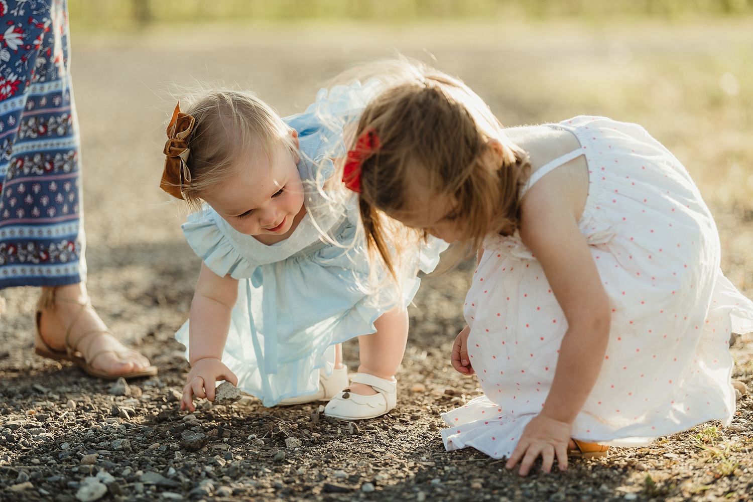 Two small children in white dresses exploring something interesting on the ground together.