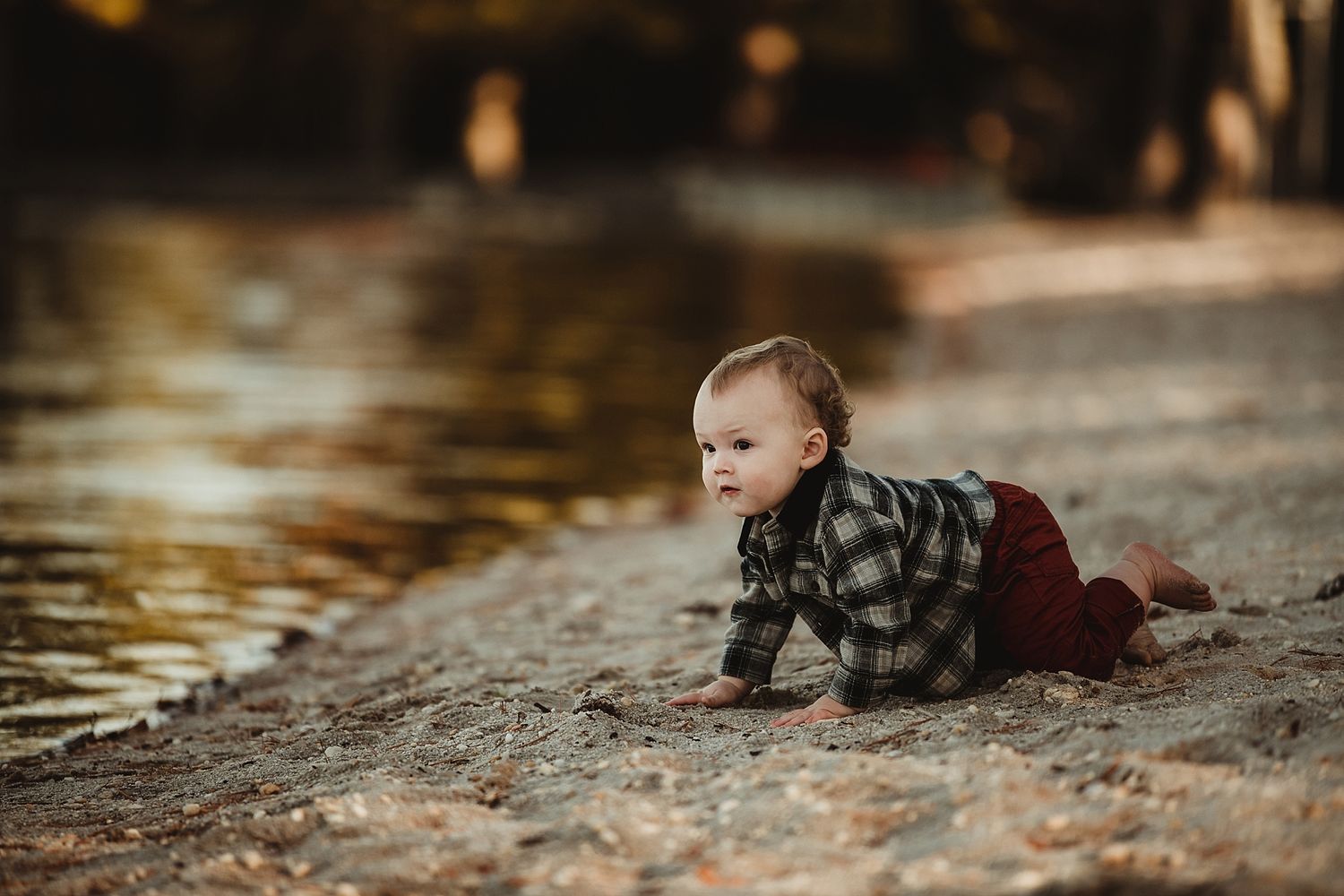 Small child in plaid shirt crawling near water's edge during golden hour.