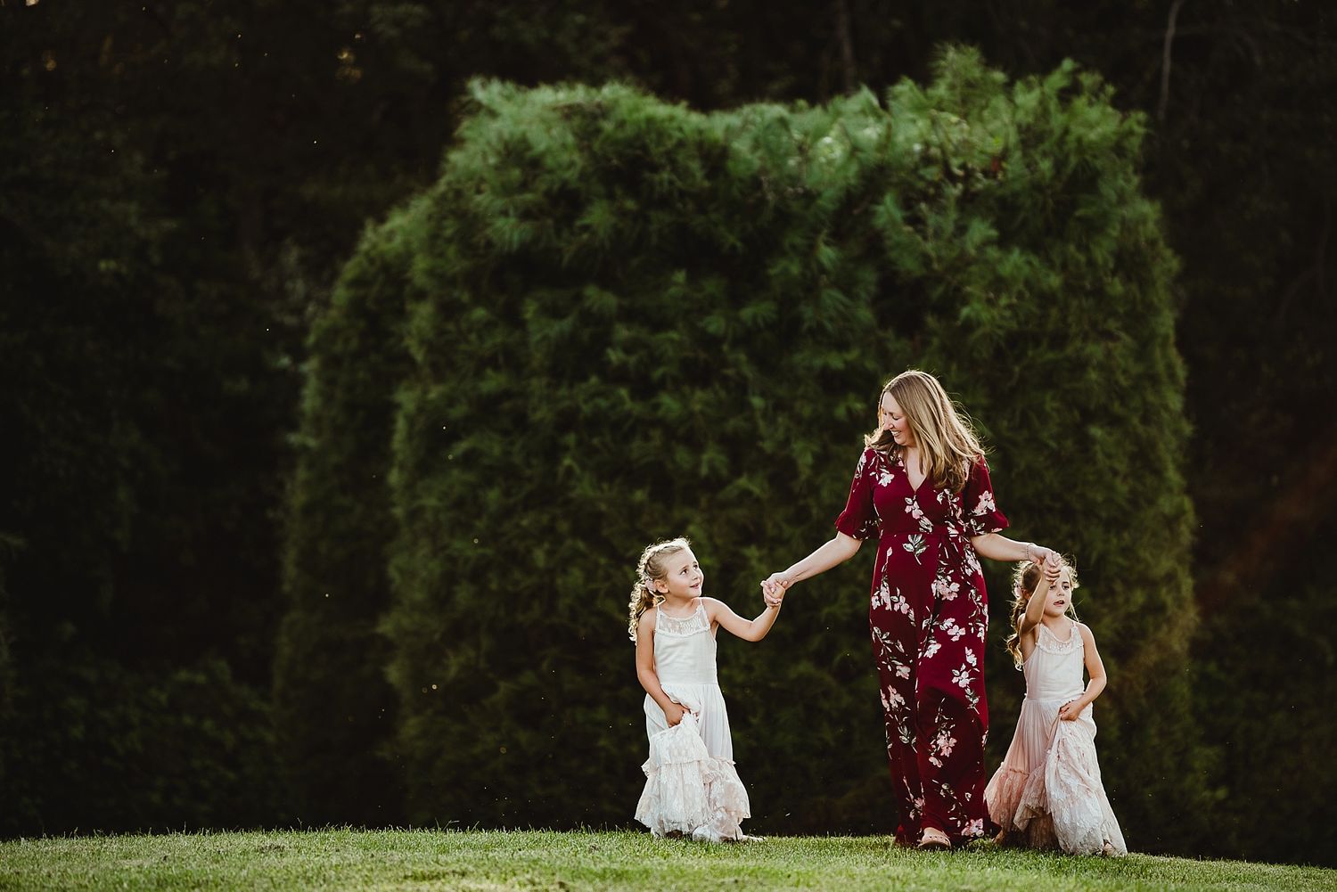 A figure in a burgundy dress dances with two children in white dresses against a backdrop of lush green hedges.