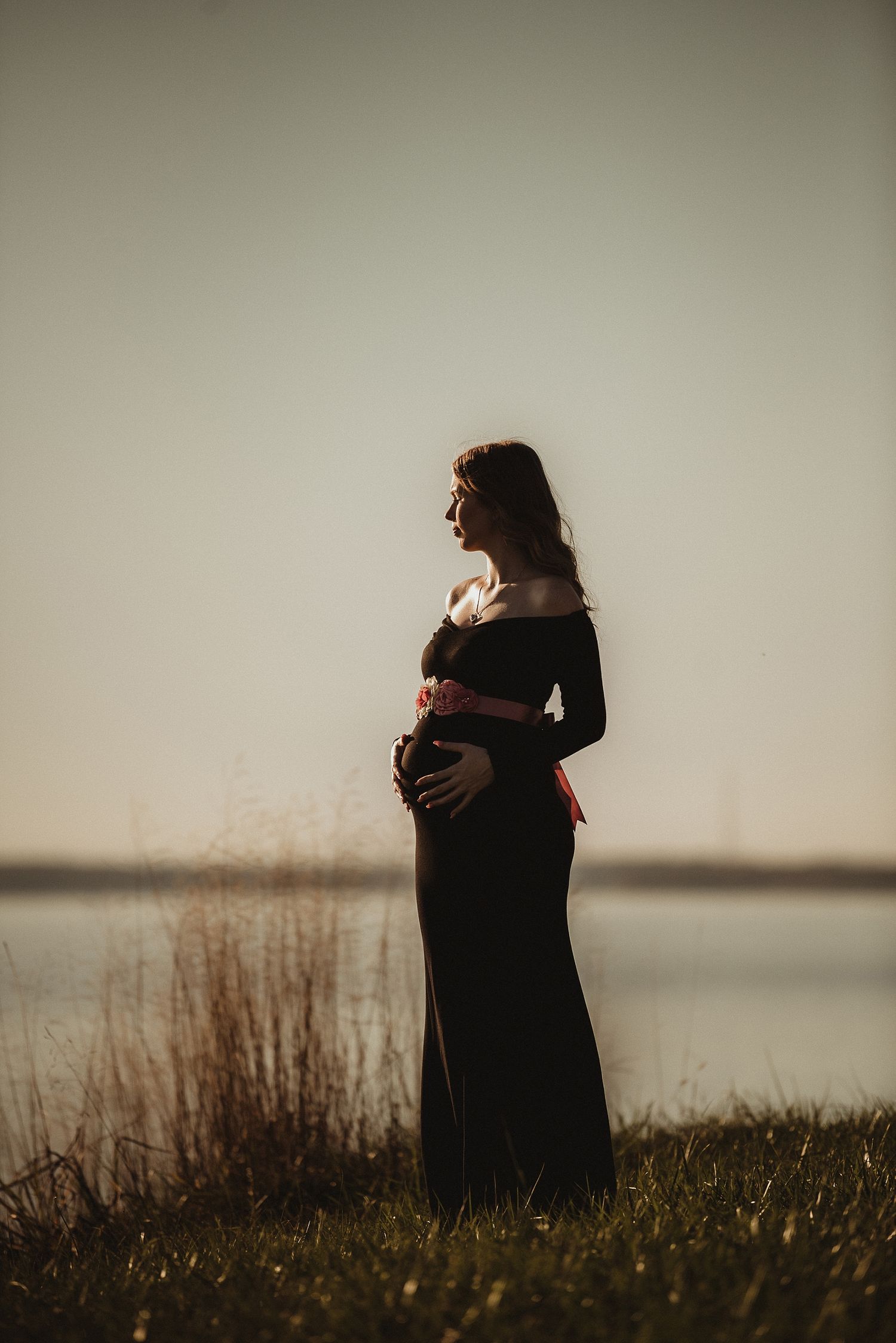 Silhouette of pregnant woman in black dress against sunset by water.