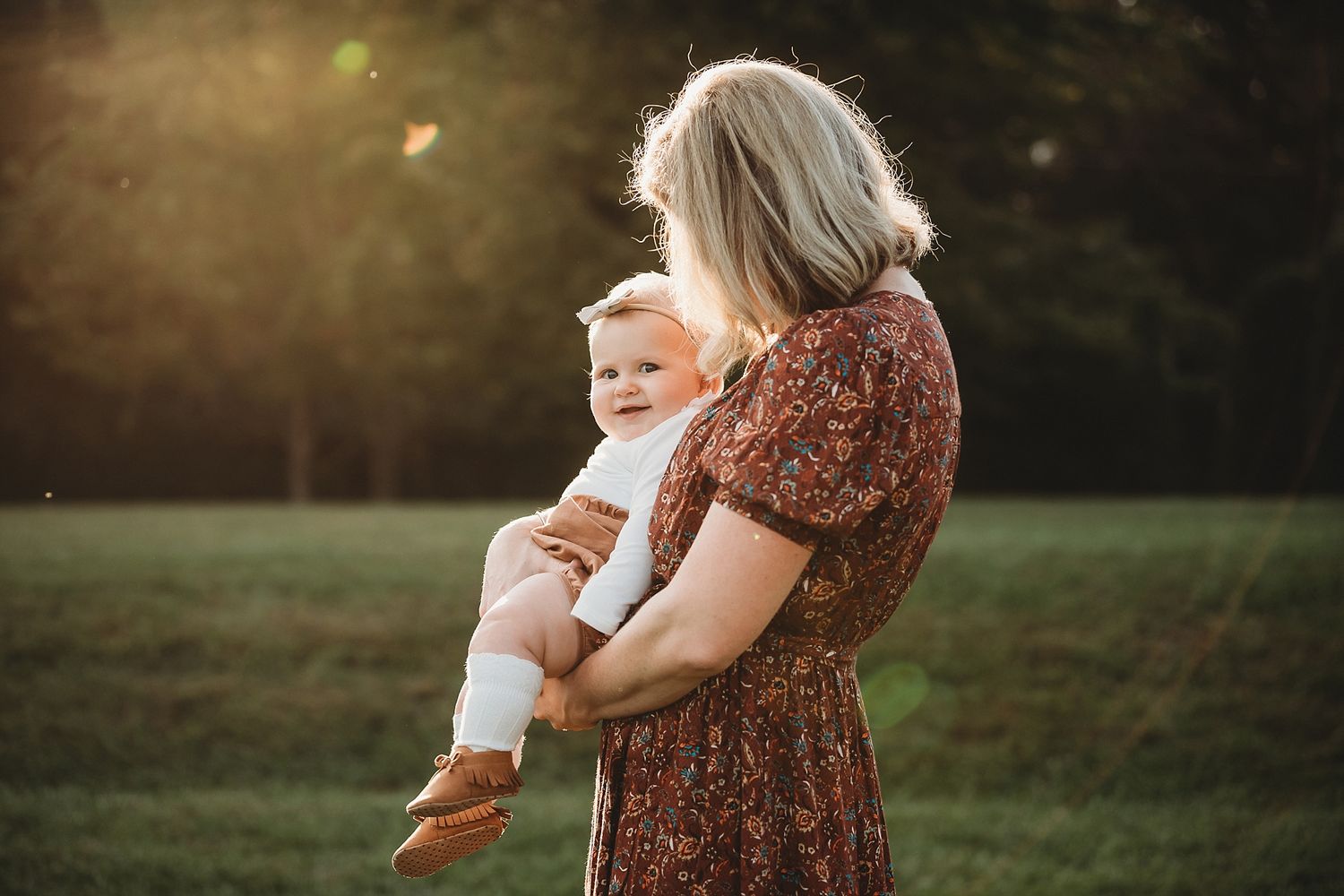Mother in floral dress holding baby during golden hour in a grassy field.