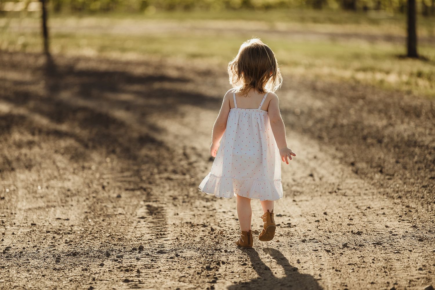 Little girl in white dress walking on dirt path during sunset.