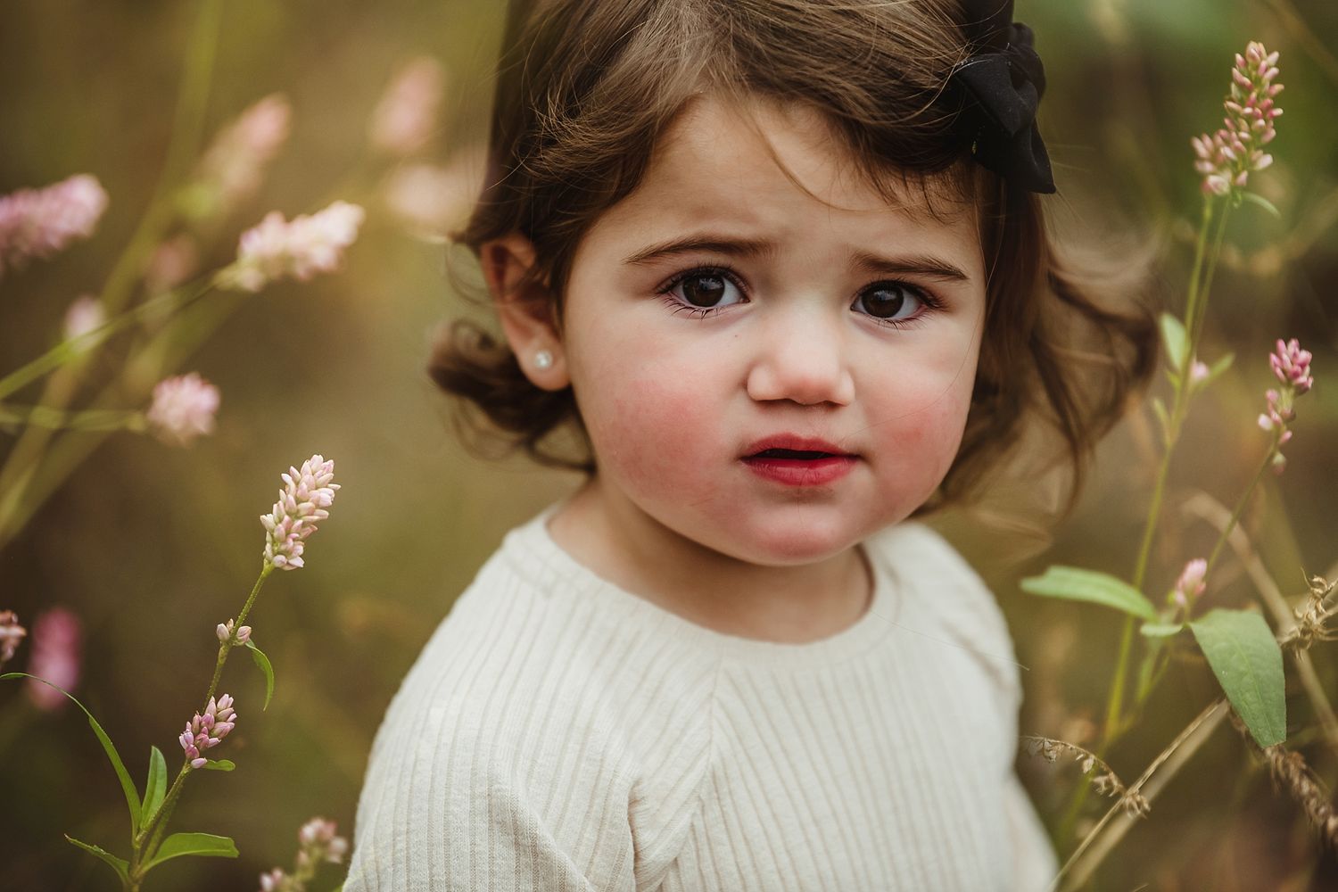 Young child in white sweater amid soft pink wildflowers in garden setting.