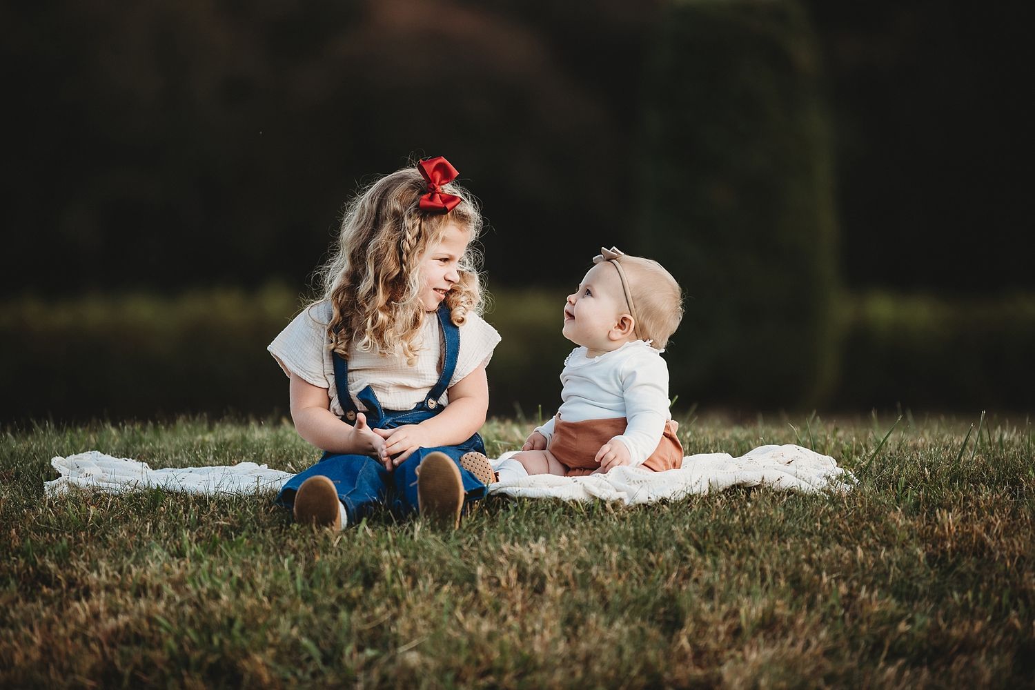 Two children sitting on white blanket outdoors in classic storybook-style portrait.