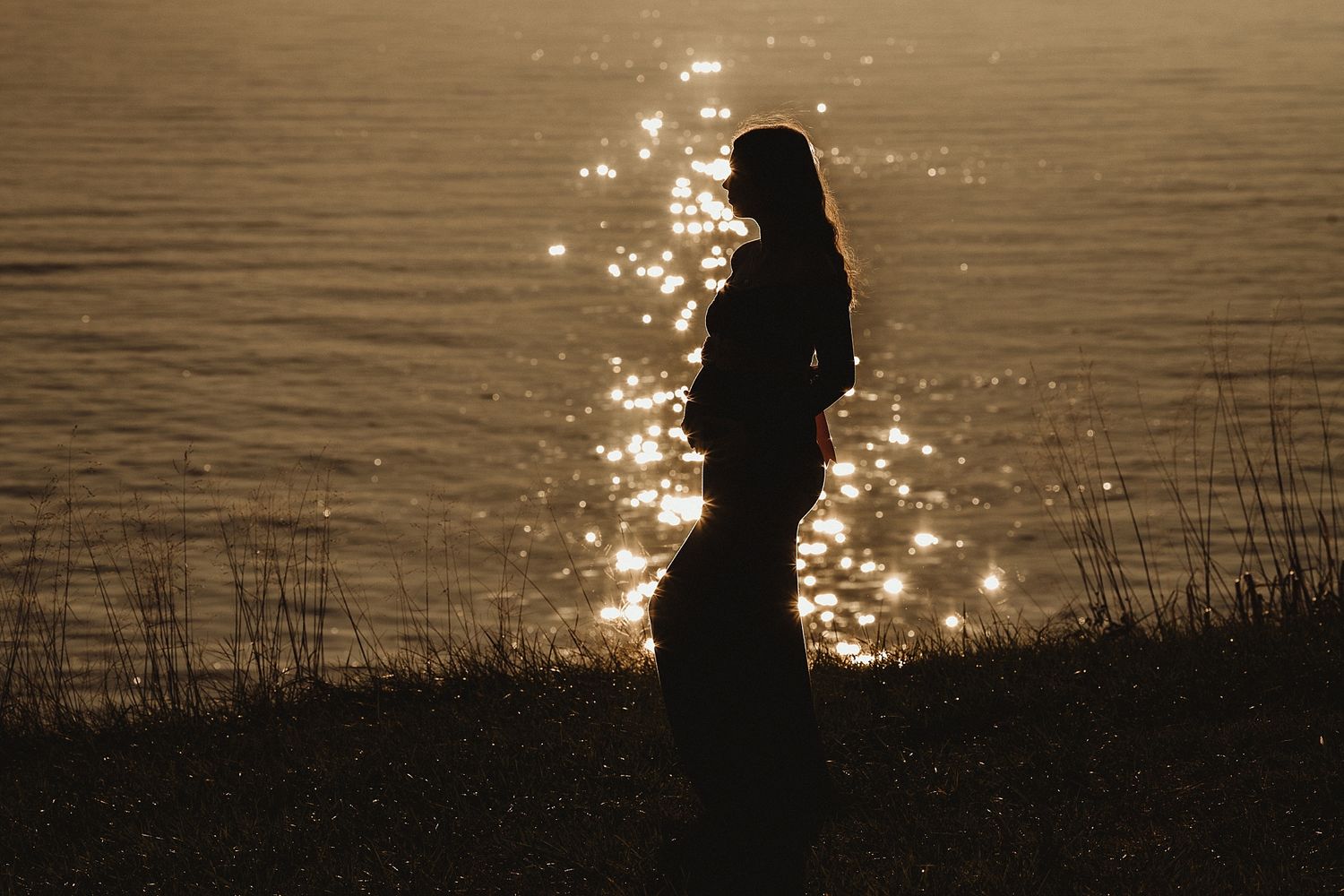 Silhouette of person standing by water at sunset with sparkling light reflections.