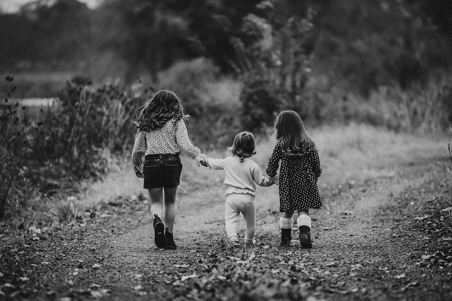 Three children walking together hand in hand on a dirt path in black and white.