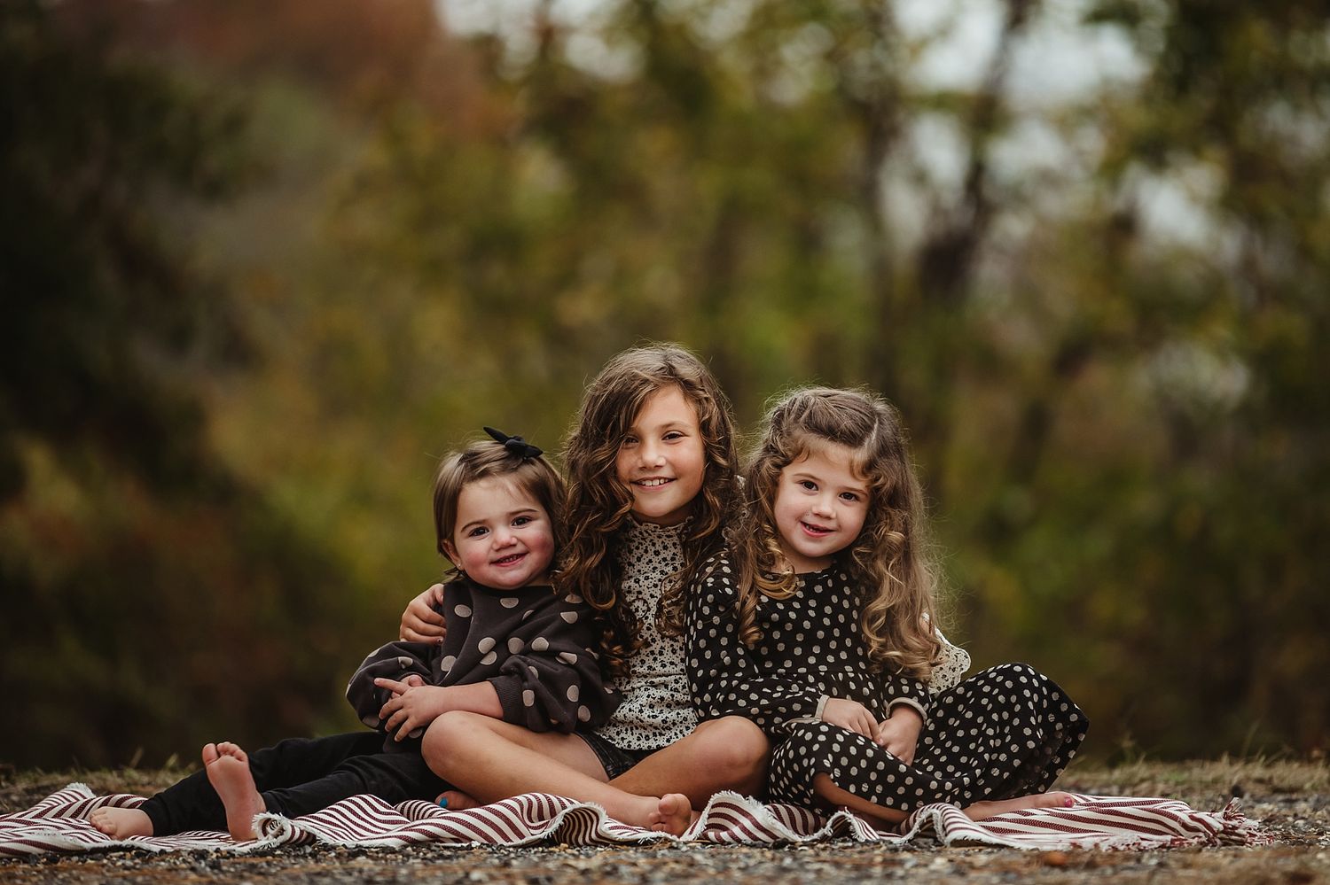 Three young children sitting together on a blanket outdoors during an autumn photoshoot.