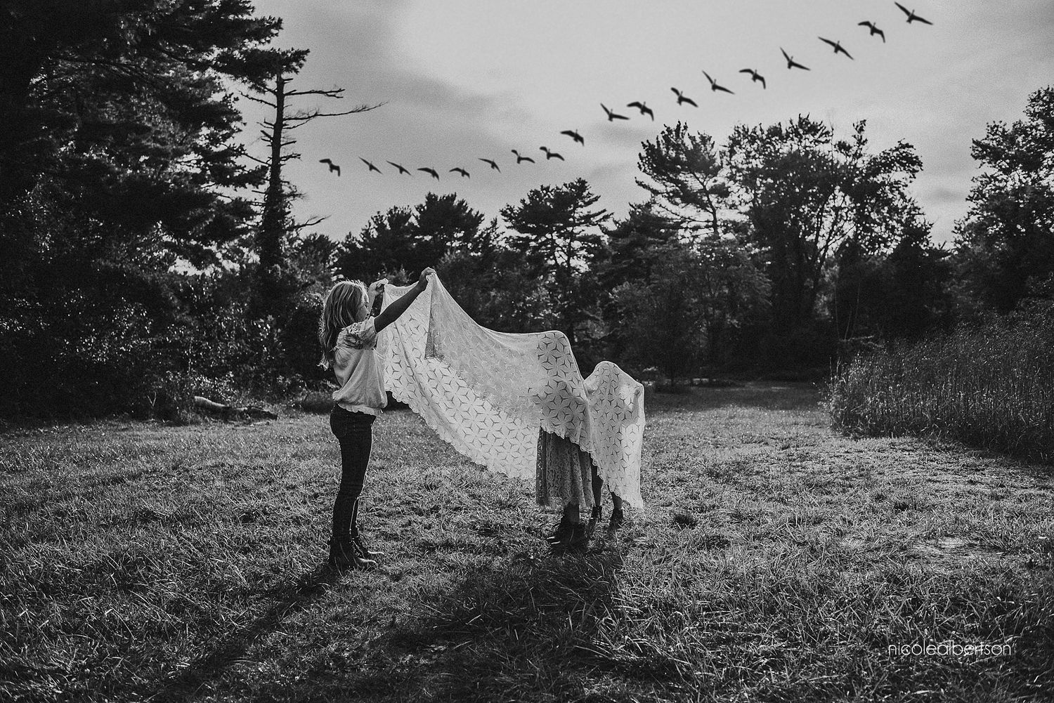 Couple holding white sheet in field with birds flying overhead in black and white.