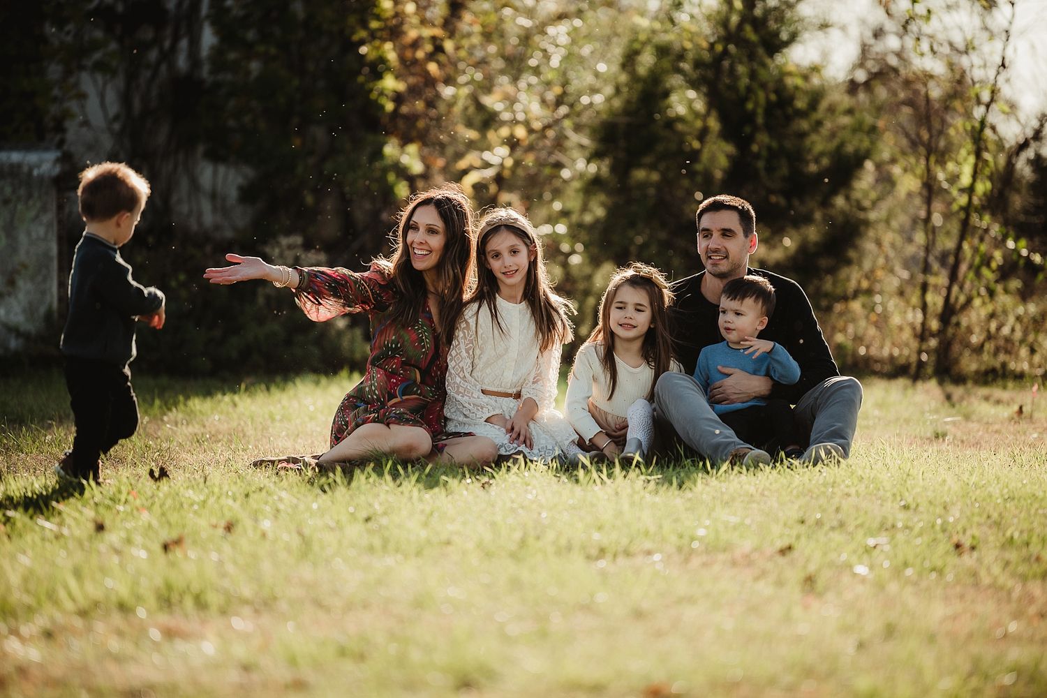 A family enjoys a playful outdoor moment on grass with sunlight filtering through trees.