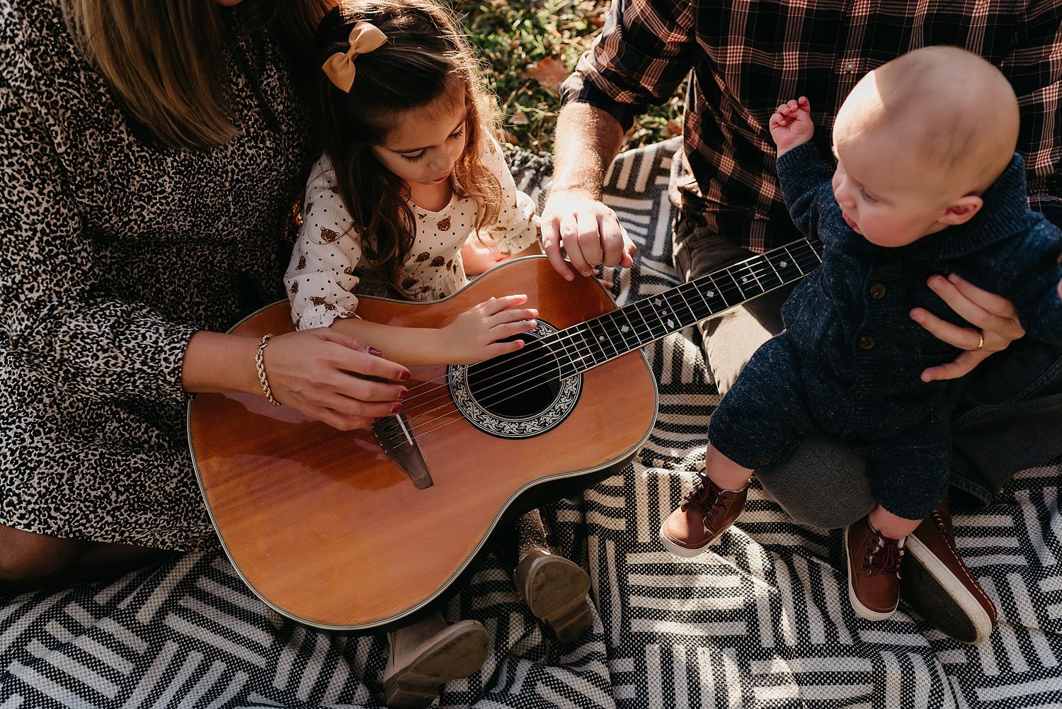 Family gathering on geometric blanket playing acoustic guitar together outdoors.