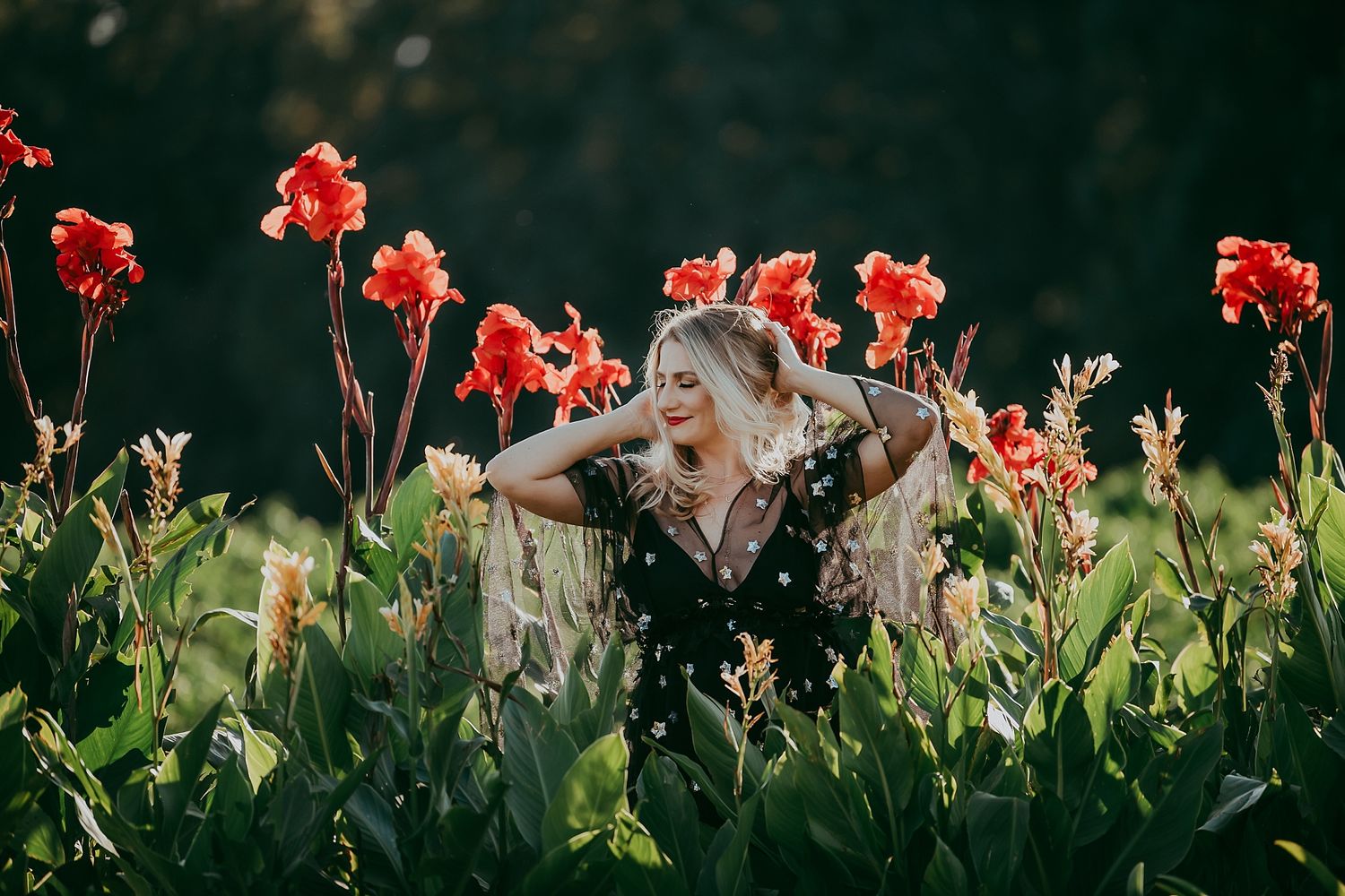 Woman joyfully ducking behind vibrant red canna lilies in a garden during golden hour light.