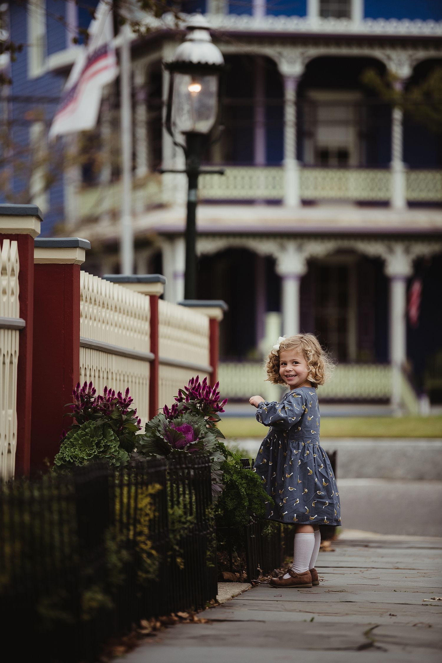 Young child in dress stands by vintage lamp post near Victorian-style house with wraparound porch.