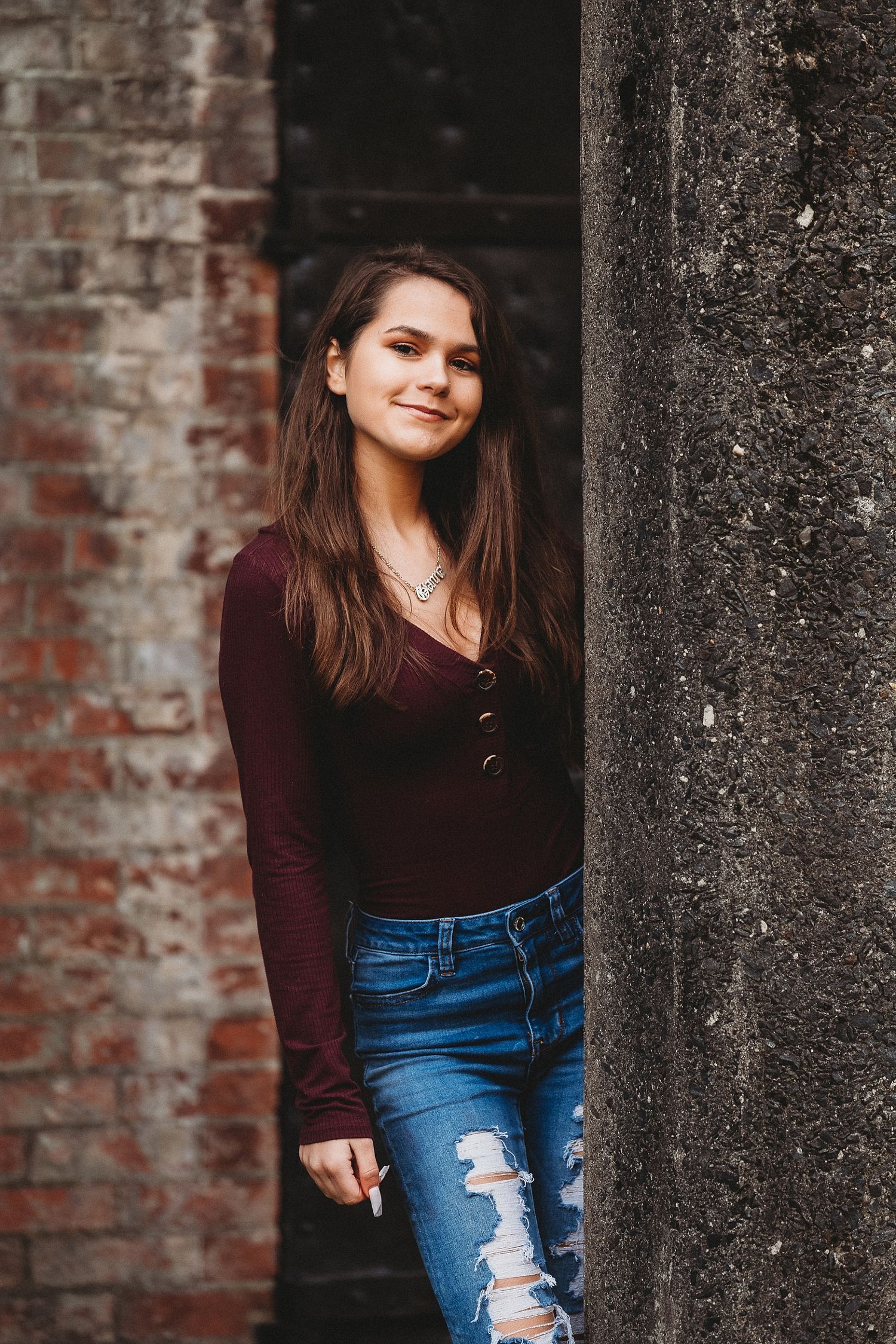 Portrait against rustic brick wall wearing burgundy top and distressed jeans.
