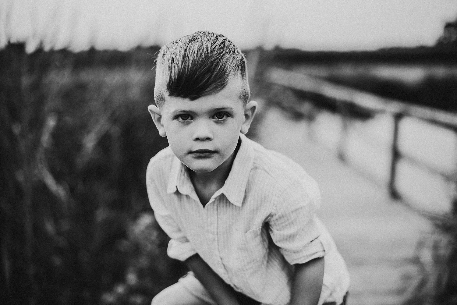 A black and white portrait of a young child leaning forward on a wooden boardwalk.