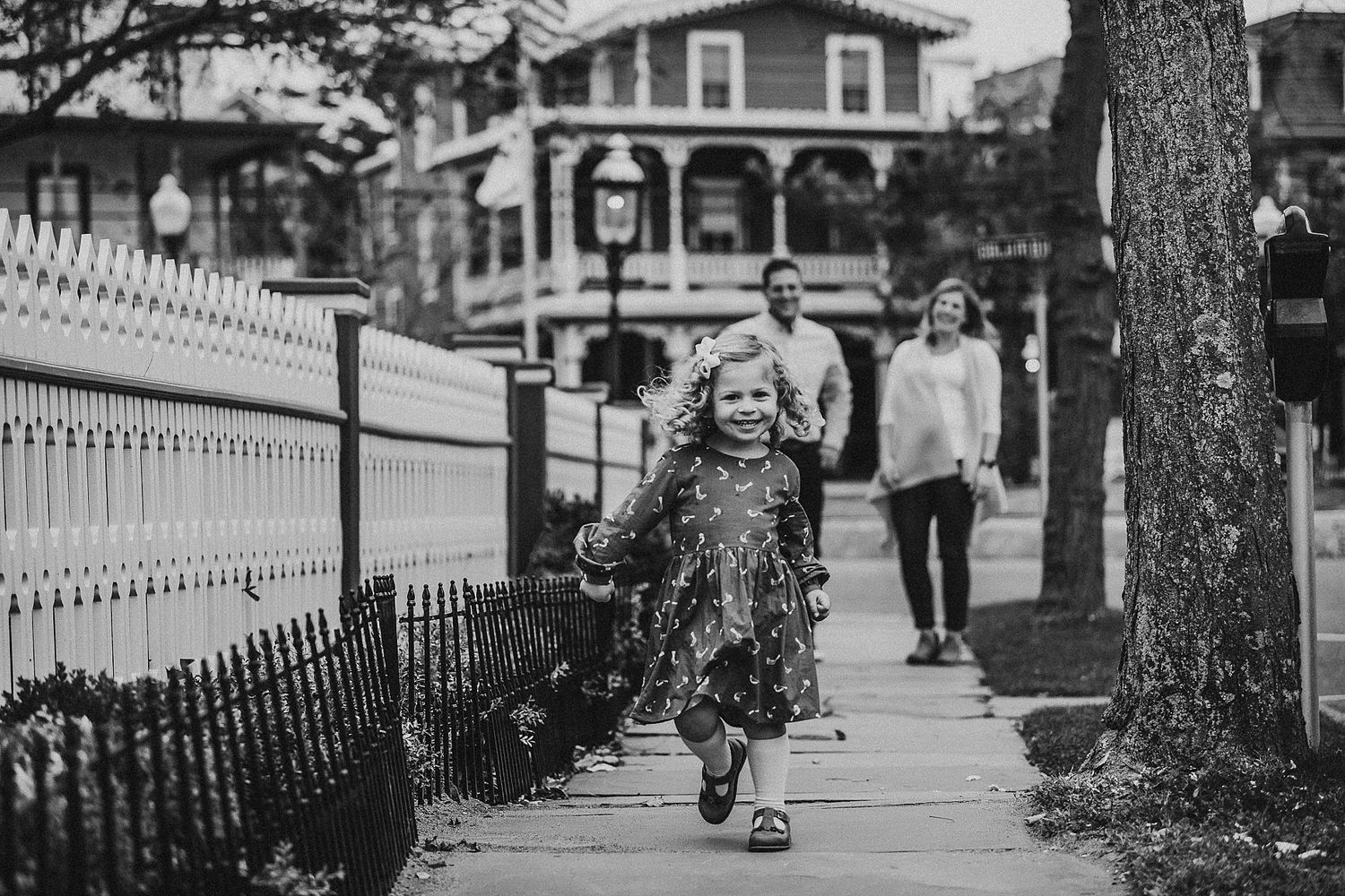 A child runs joyfully along a sidewalk with a white picket fence in black and white.