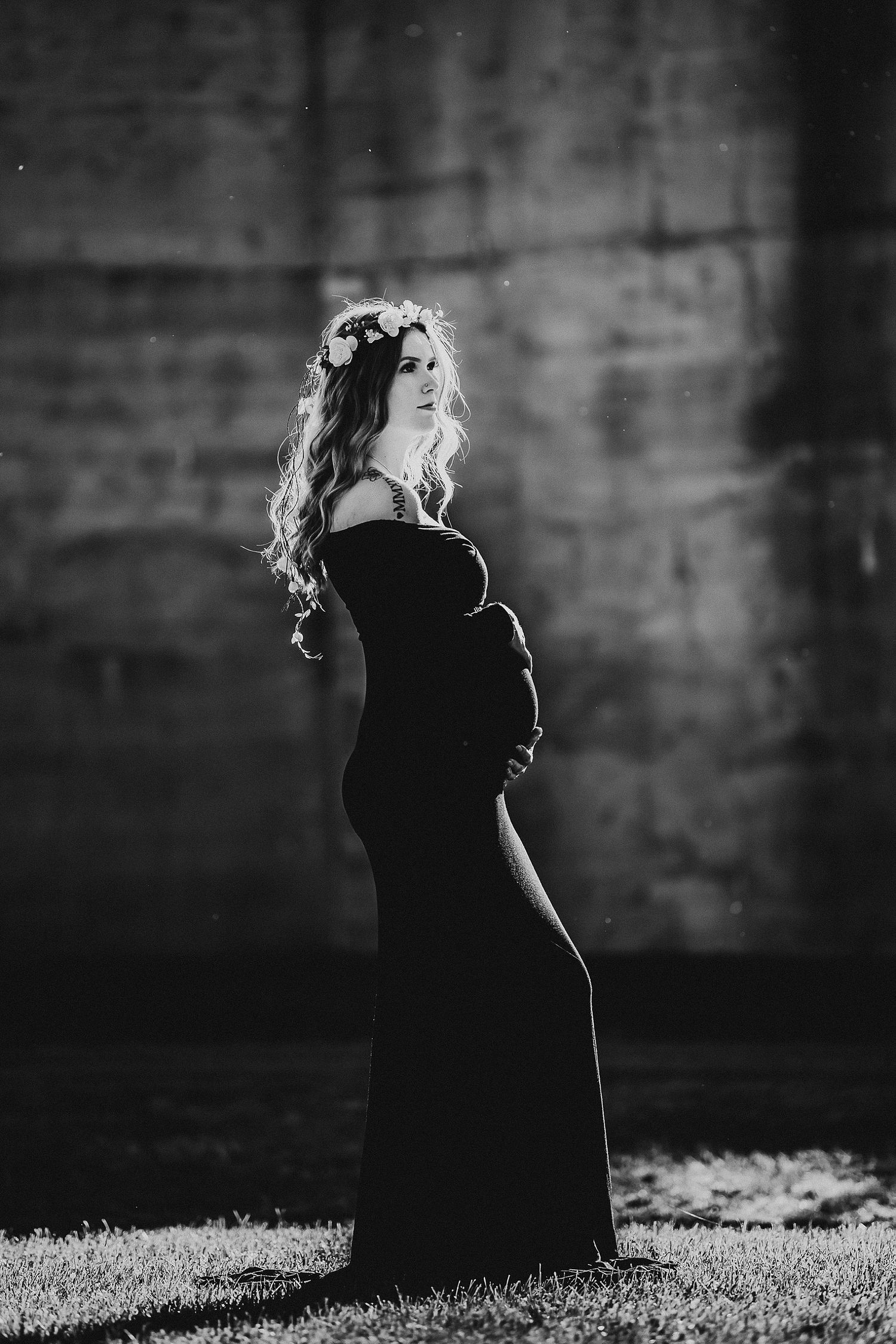 A silhouette of an expectant mother wearing a black dress and flower crown against a textured wall.