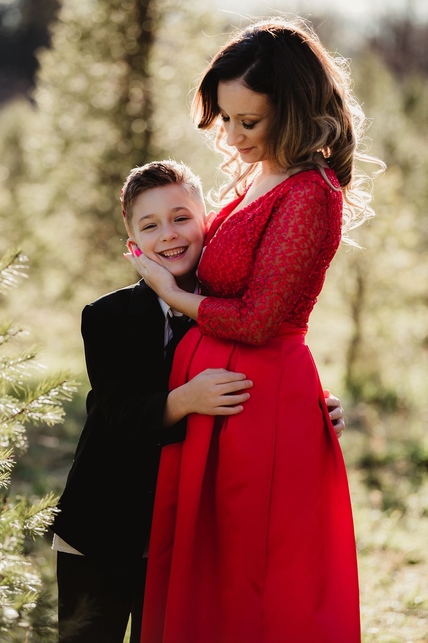 A touching maternity moment captured in a red lace dress against a sunlit background.
