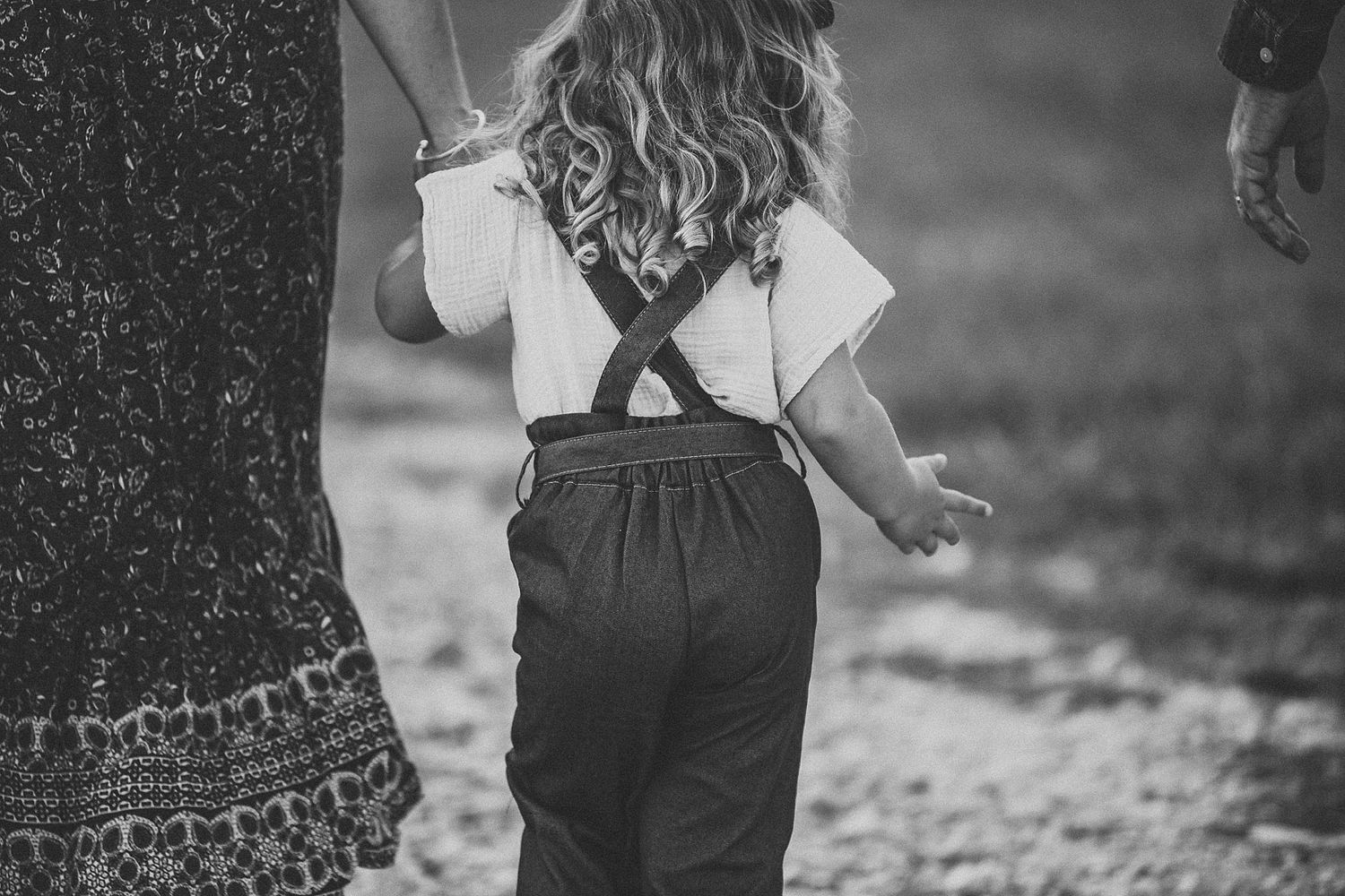 Back view of a small child in overalls walking while holding hands with someone in a patterned dress.