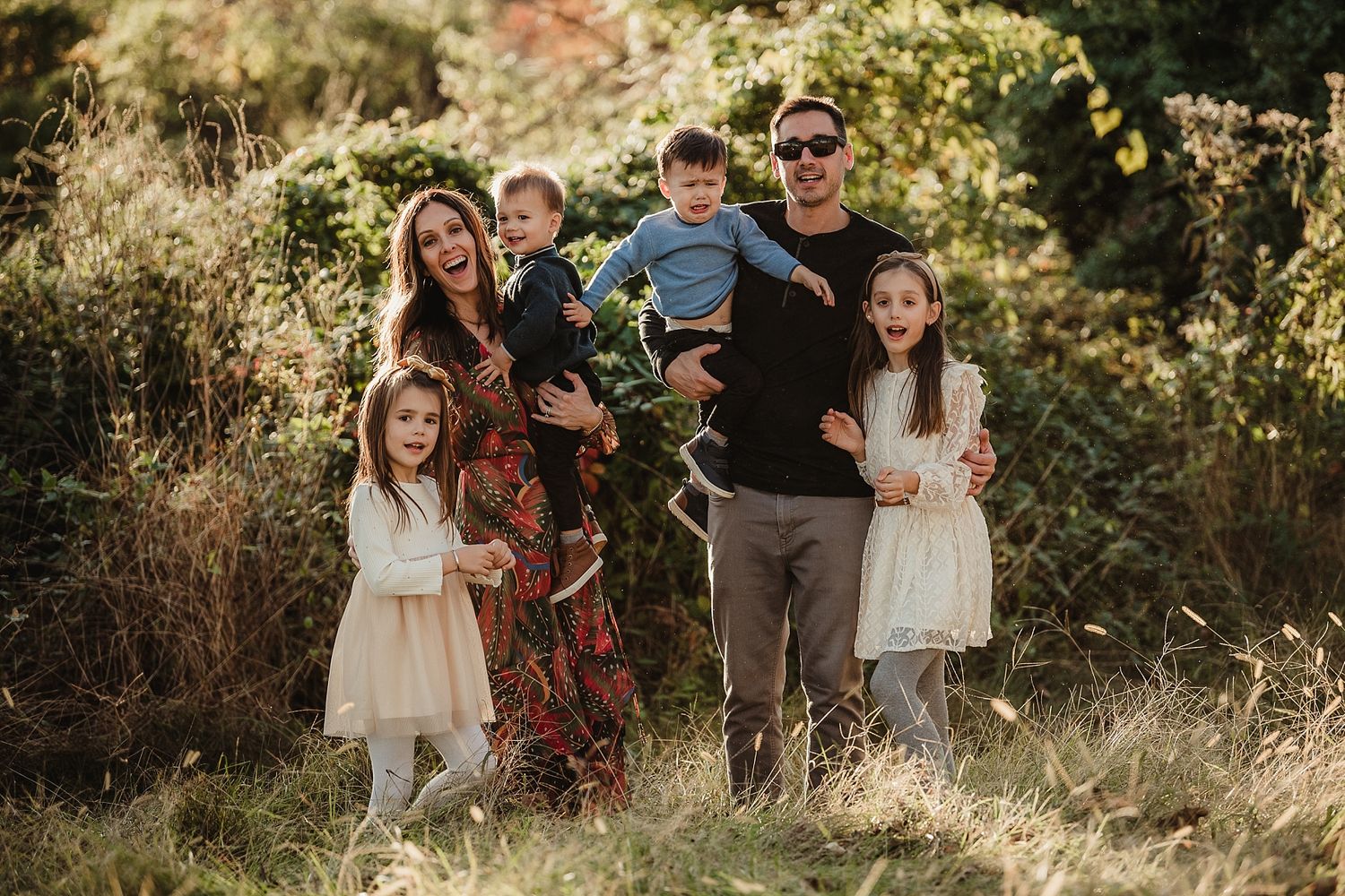 Happy family of six poses together in a sunlit field during golden hour.