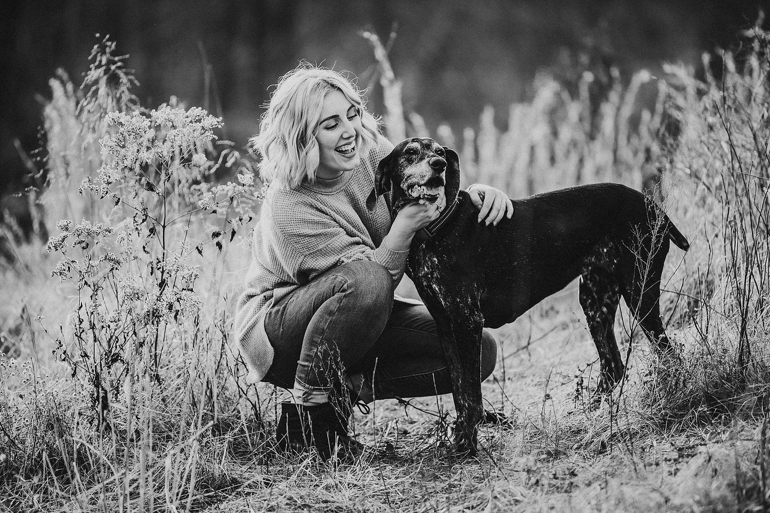 Black and white portrait of someone crouching down to pet their black dog in a field.