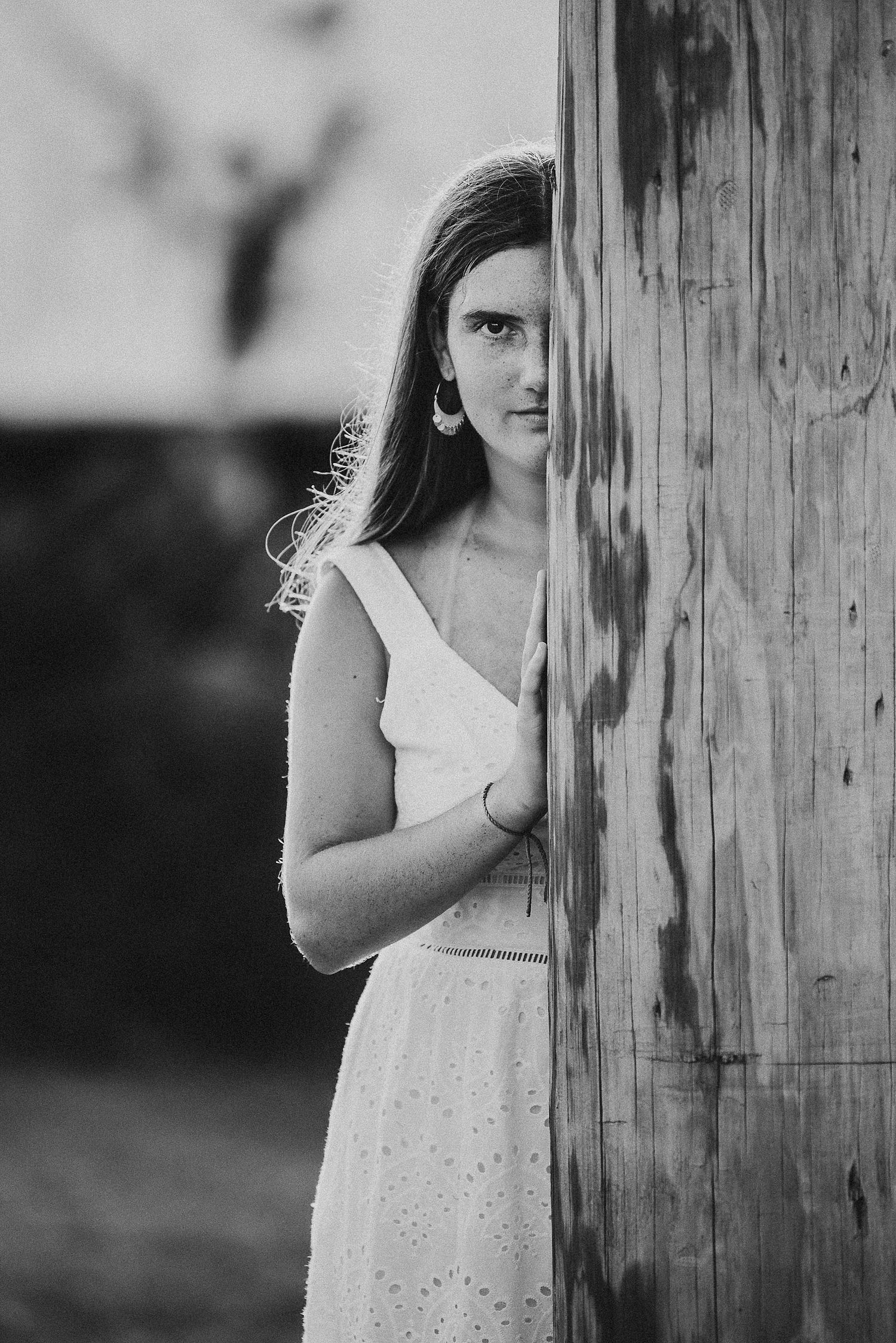 A black and white artistic portrait of someone peeking from behind a wooden post.
